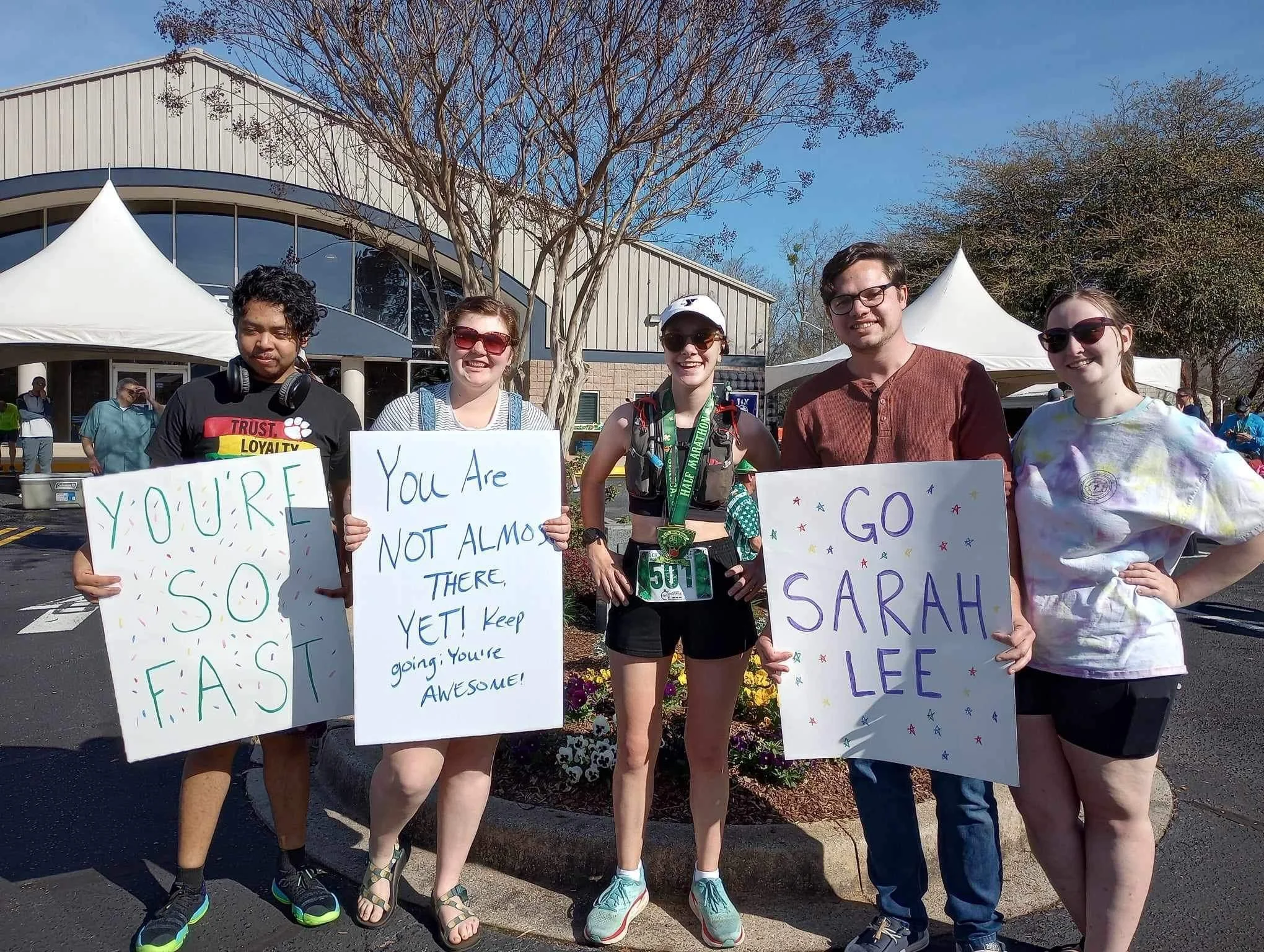 Five young adults standing outside near a building with trees in the background, holding signs with encouraging messages and a race bib, suggesting they are at a running event or marathon on a sunny day.