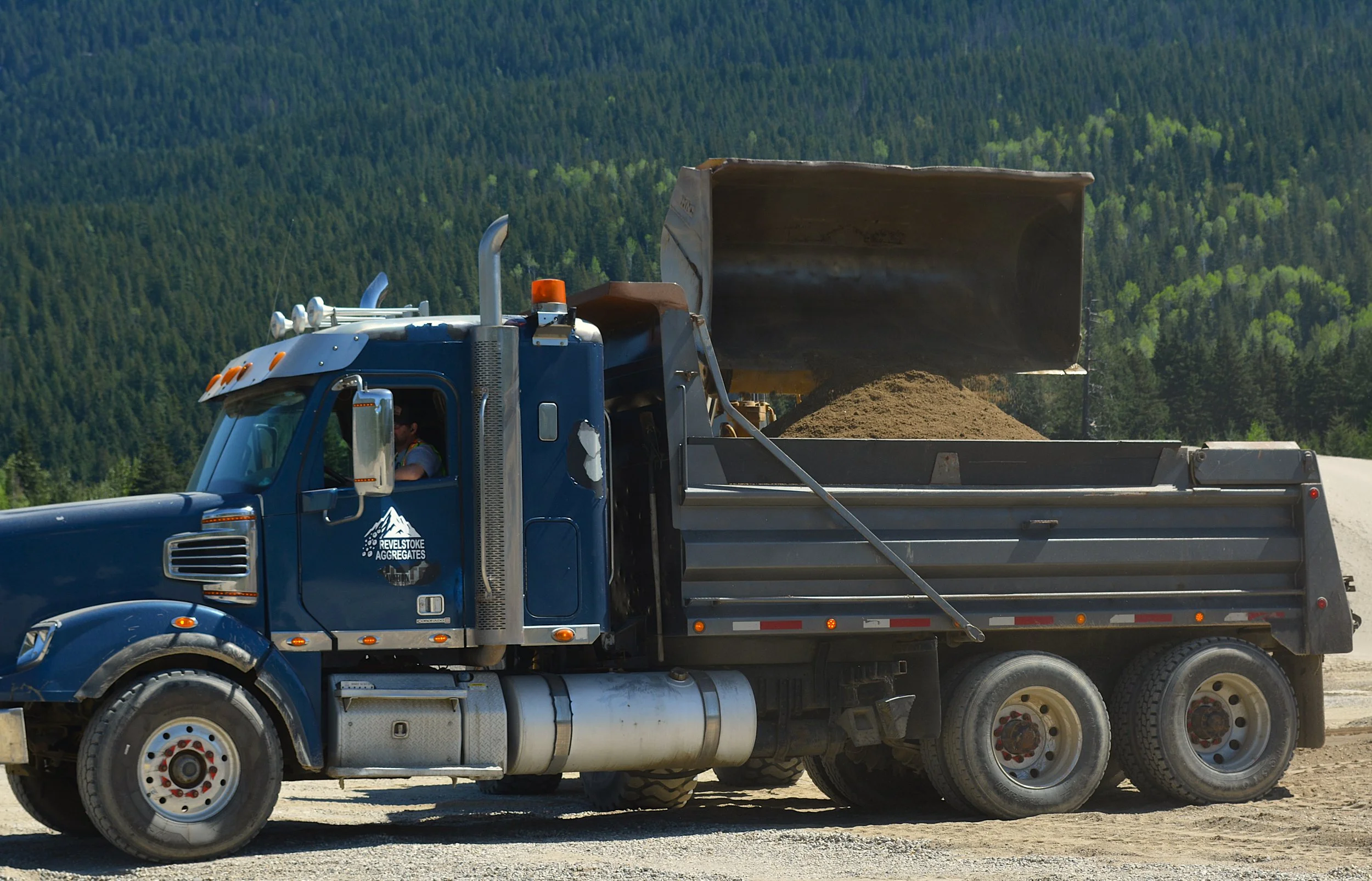 Blue dump truck with dirt in the bed driving on a gravel road, with a forested mountain range in the background.