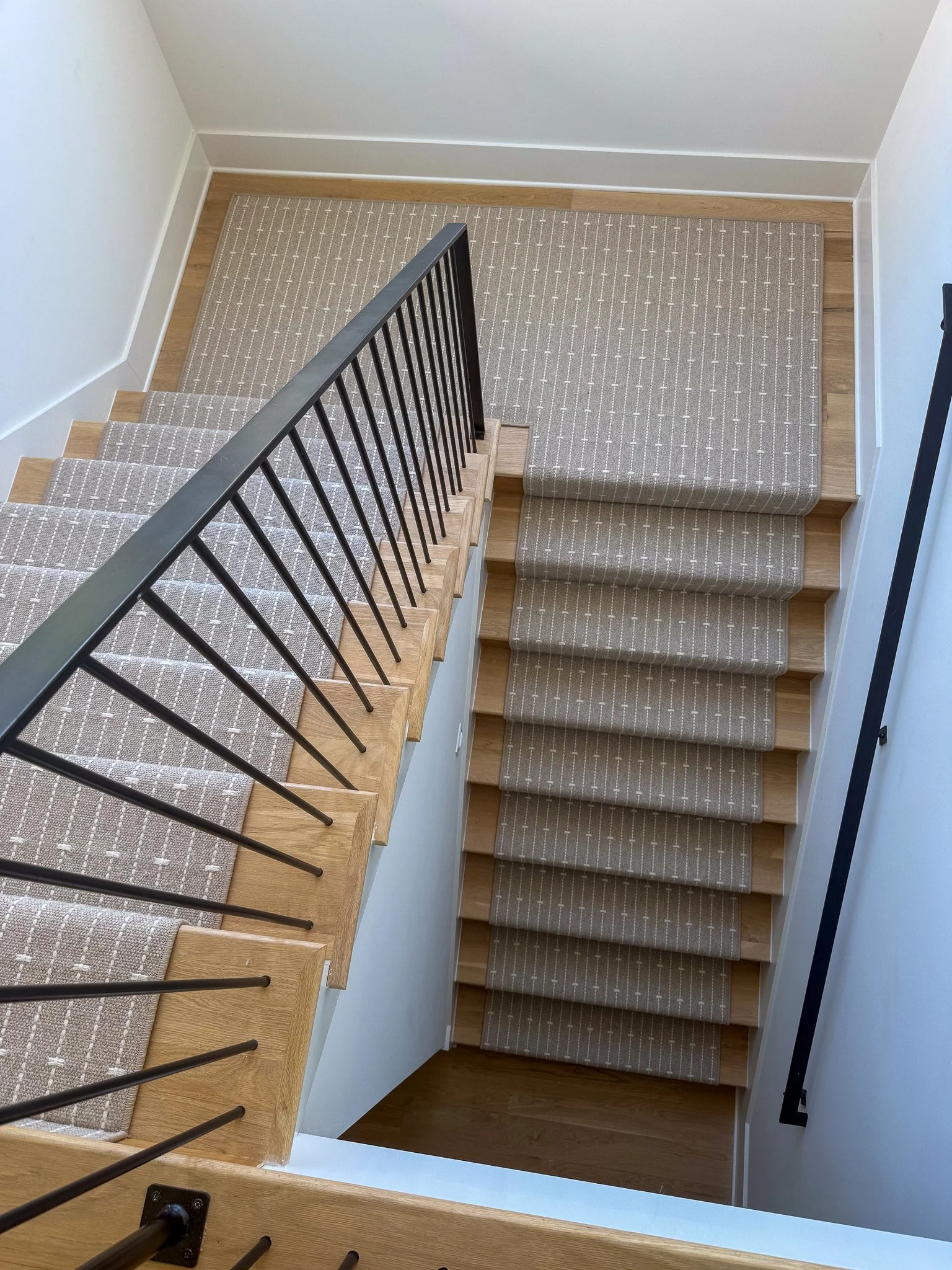 Top view of a staircase with wood steps, a beige runner with white pattern, black metal handrails, and white walls.