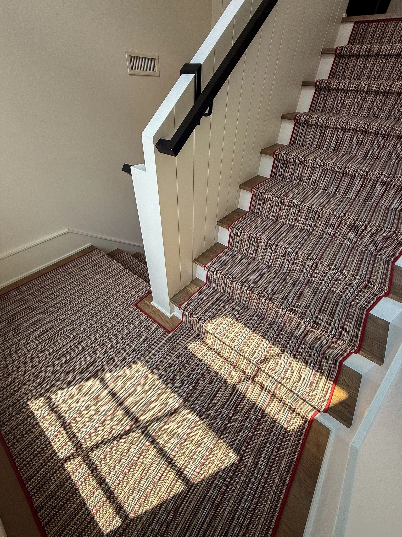 View of a staircase with multicolored striped carpet, white and black handrails, and beige walls. Sunlight creates shadow patterns on the stairs and carpet.