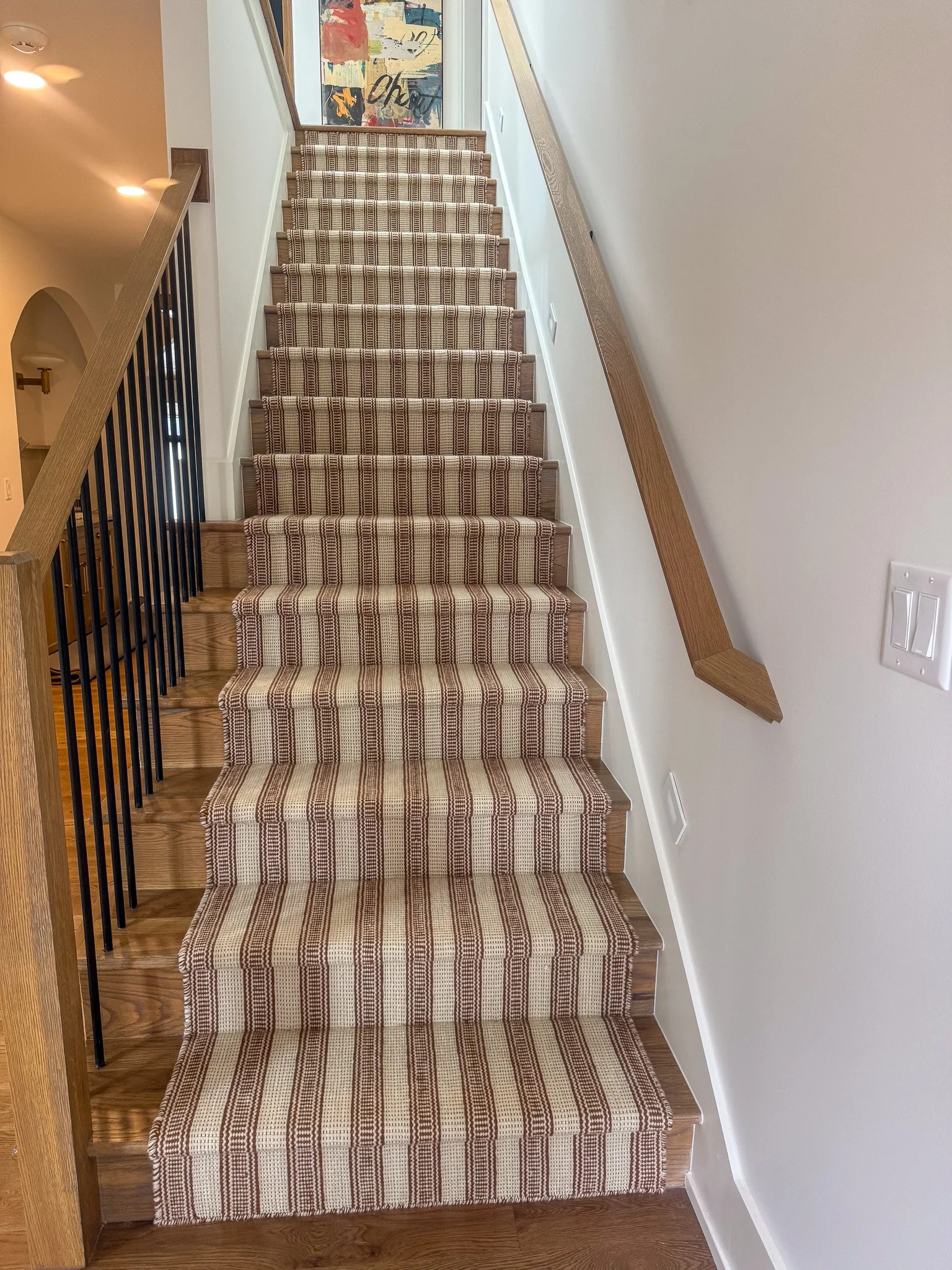 Carpeted wooden staircase with a patterned runner, beige and brown stripes, leading to an upper floor with a colorful abstract painting on the wall in the background.