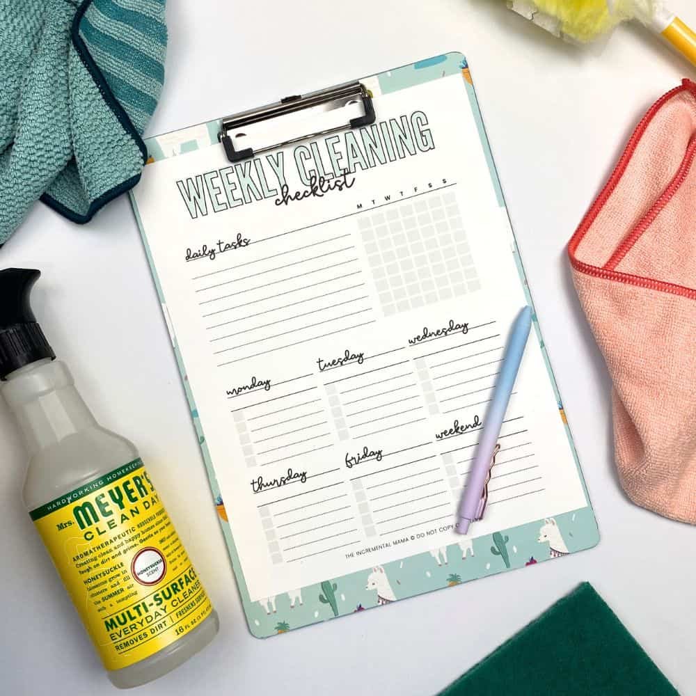 A weekly cleaning checklist on a clipboard, with cleaning supplies surrounding it, including a spray bottle, cleaning cloths, and a pink zippered pouch.