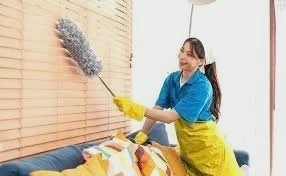 A woman wearing a blue shirt and yellow gloves cleaning a couch with a duster in a living room.