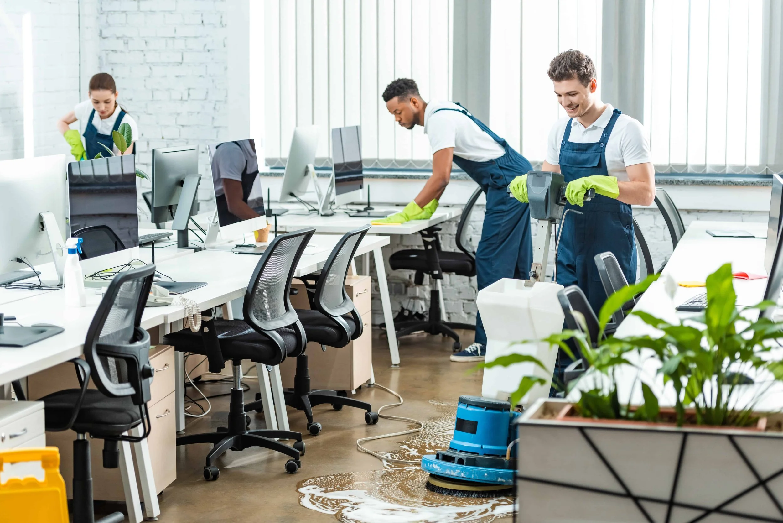 Three people cleaning an office with disinfectant and cleaning equipment, with computers, chairs, and plants visible.