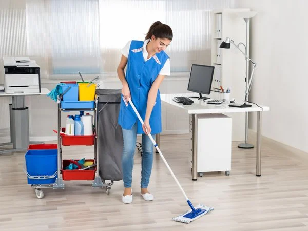 A woman in a blue uniform cleaning a wooden floor with a mop in an office setting.