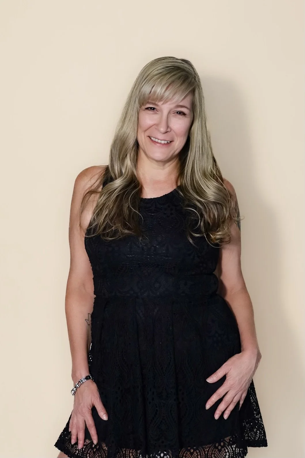 A woman with long, wavy blonde hair wearing a sleeveless black lace dress, standing against a gray background.