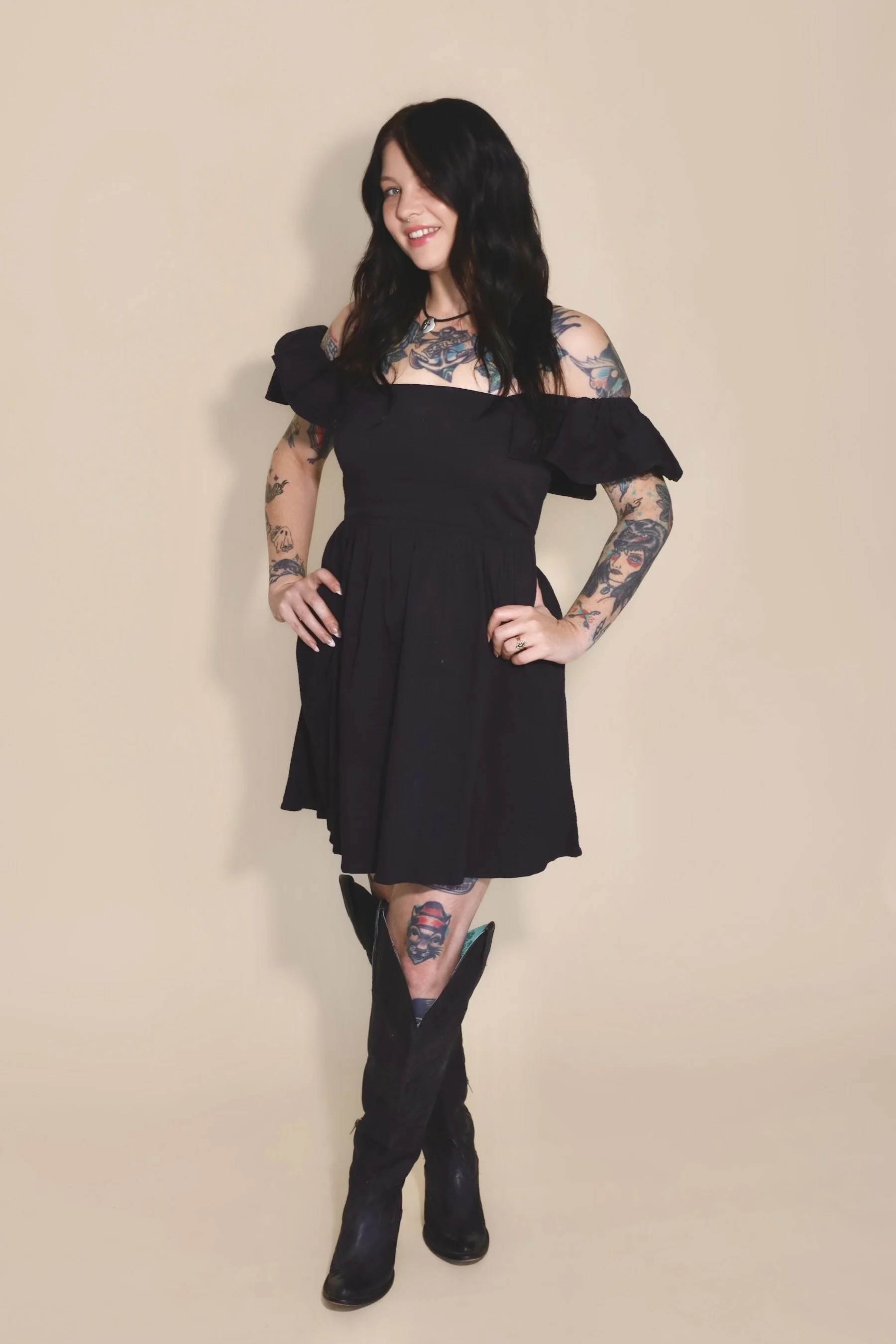 Portrait of a young woman with black hair, tattoos on her arms, wearing a black dress and a necklace with a cross, standing against a dark background.