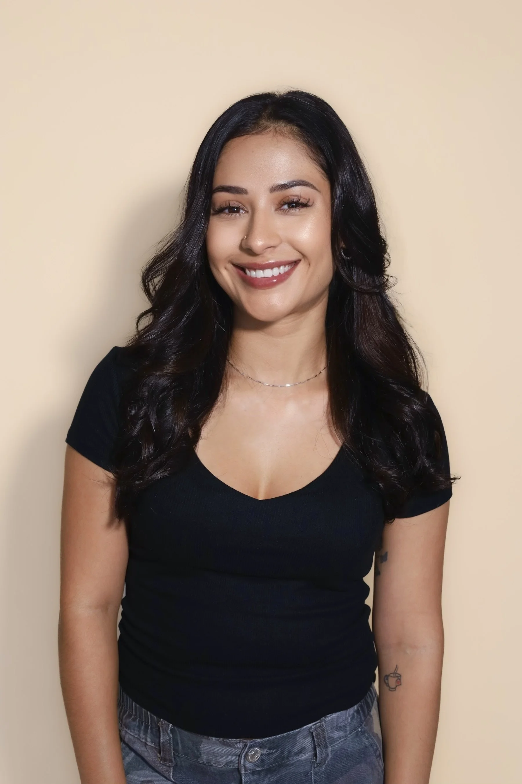 A woman with dark, wavy hair wearing a black headband and a black top with her name and credentials embroidered on it, smiling softly at the camera against a dark background.