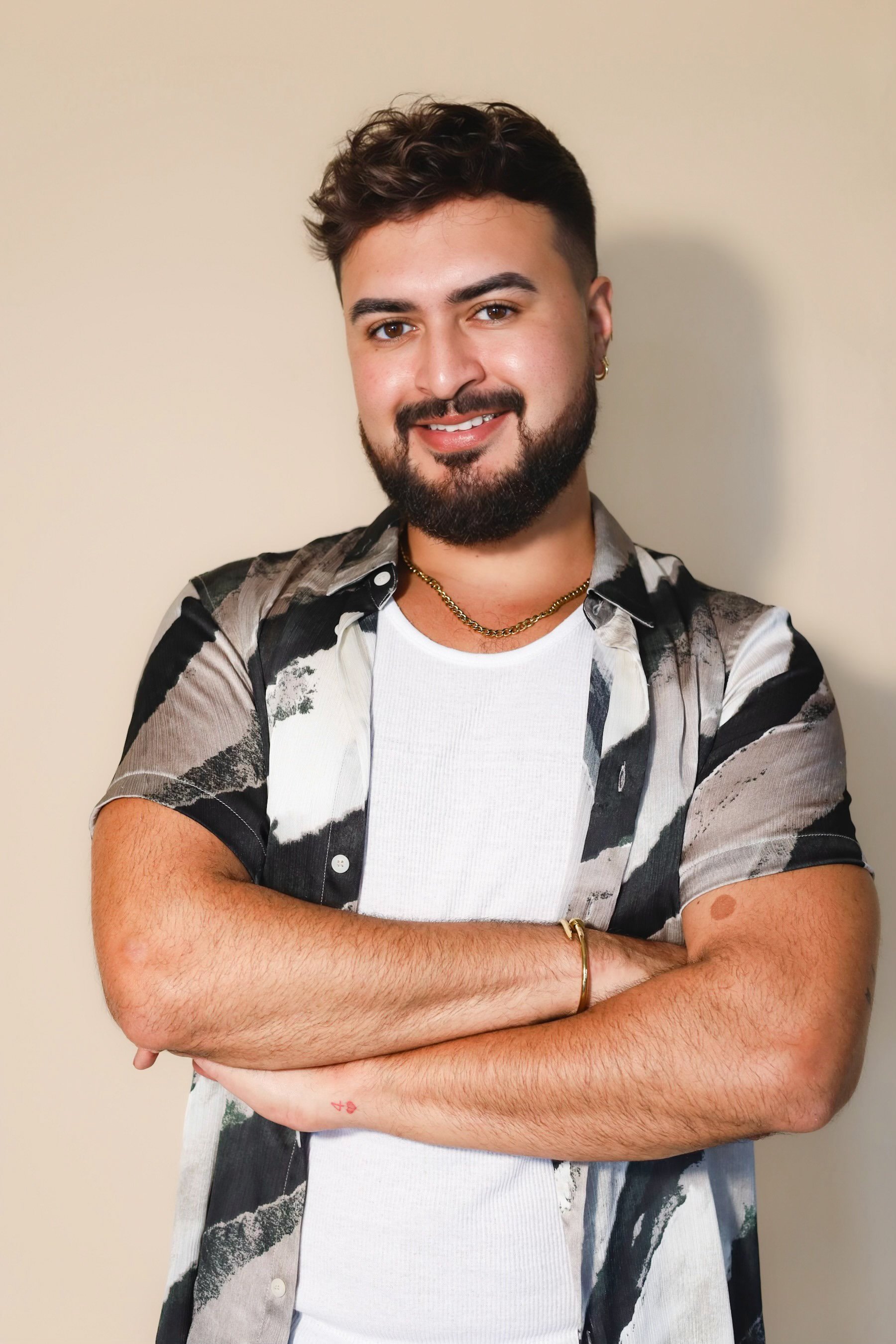 A young man with a beard and short dark hair, wearing a black polo shirt, silver necklace, bracelet, and watch, smiling with arms crossed against a dark background.