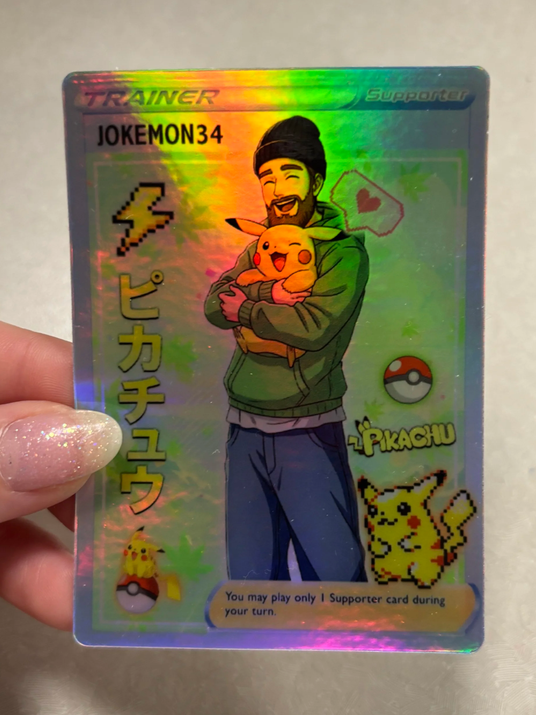 Holographic Pokémon supporter card featuring a cartoon man with a beard holding Pikachu. The card has Japanese text and Pokémon symbols, including a Poké Ball, Pikachu, and a star. The card is labeled 'JOKEMON34' and includes instruction text about p
