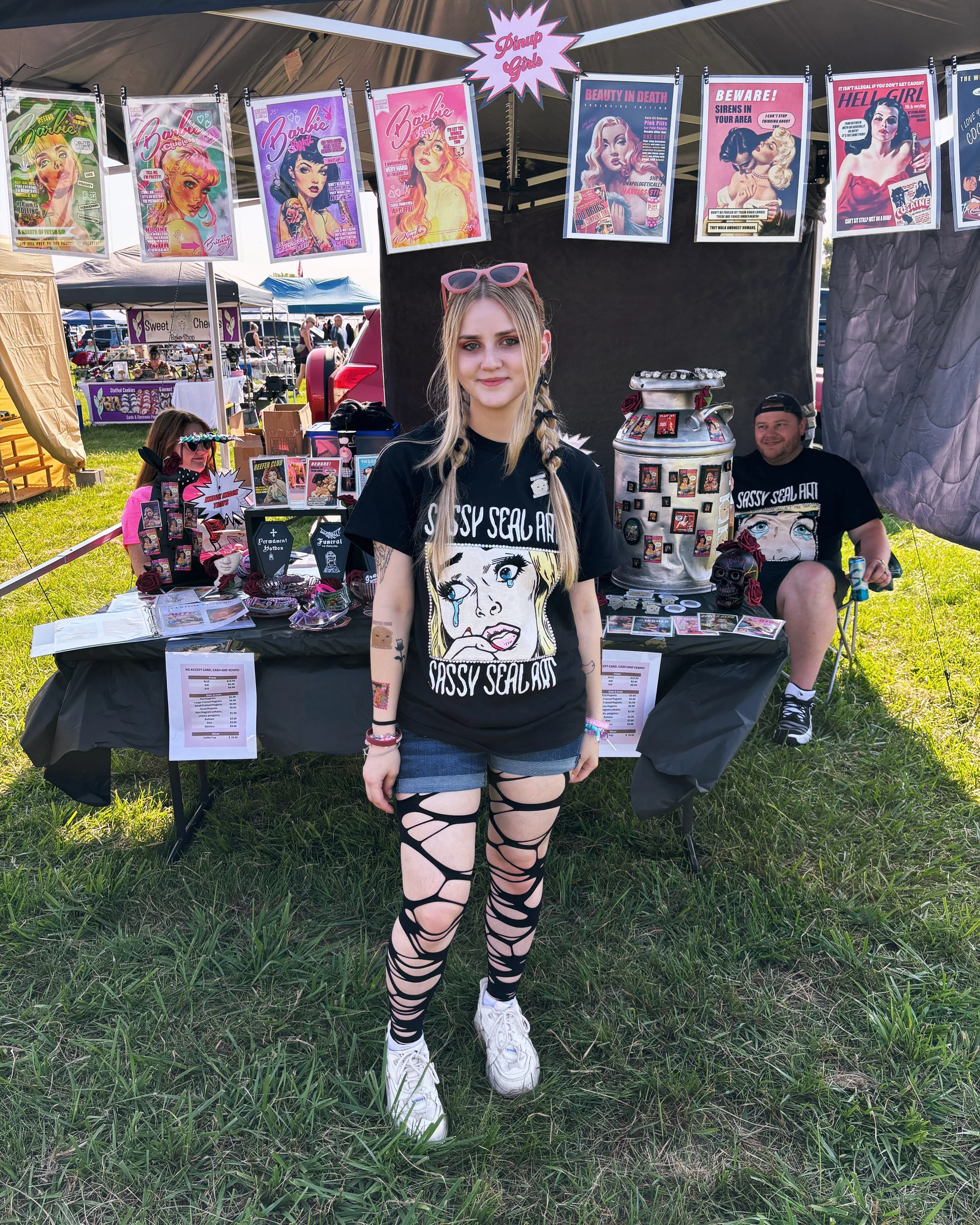 Young woman standing in front of a booth at an outdoor festival, wearing a black 'Sassy Seal Art' T-shirt with a cartoon girl on it. The booth behind her has various custom sticker designs and pinup girl posters.