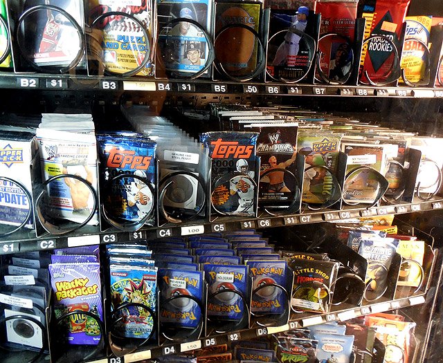 Vending machine shelf with collectible items including lenses, cards, and Pokémon trading card packs.
