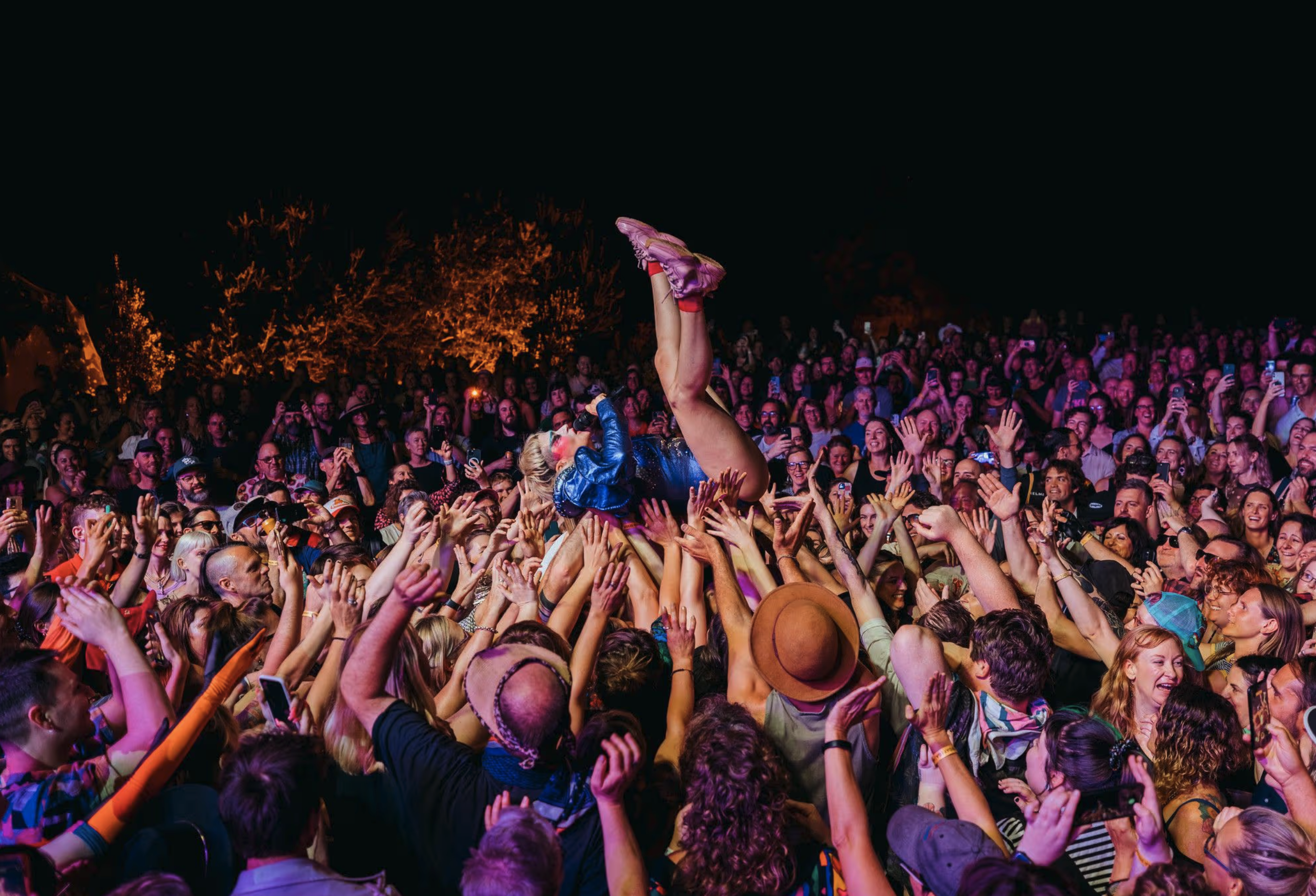 A woman crowd surf at a music festival during nighttime, surrounded by a large audience cheering and reaching out to her, with lights illuminating the scene.