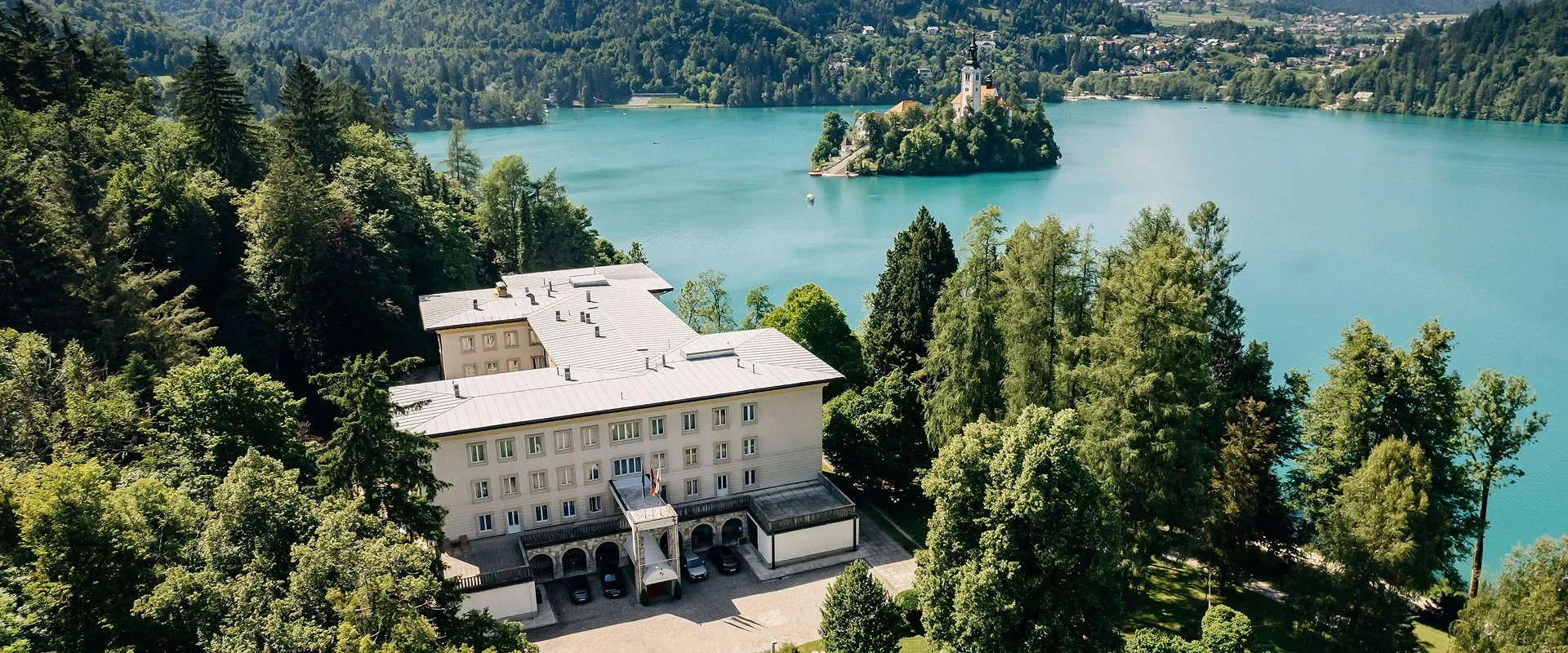 Aerial view of Lake Bled with an island featuring a church, surrounded by lush forest and mountains, and a large building with a metal roof near the lake.