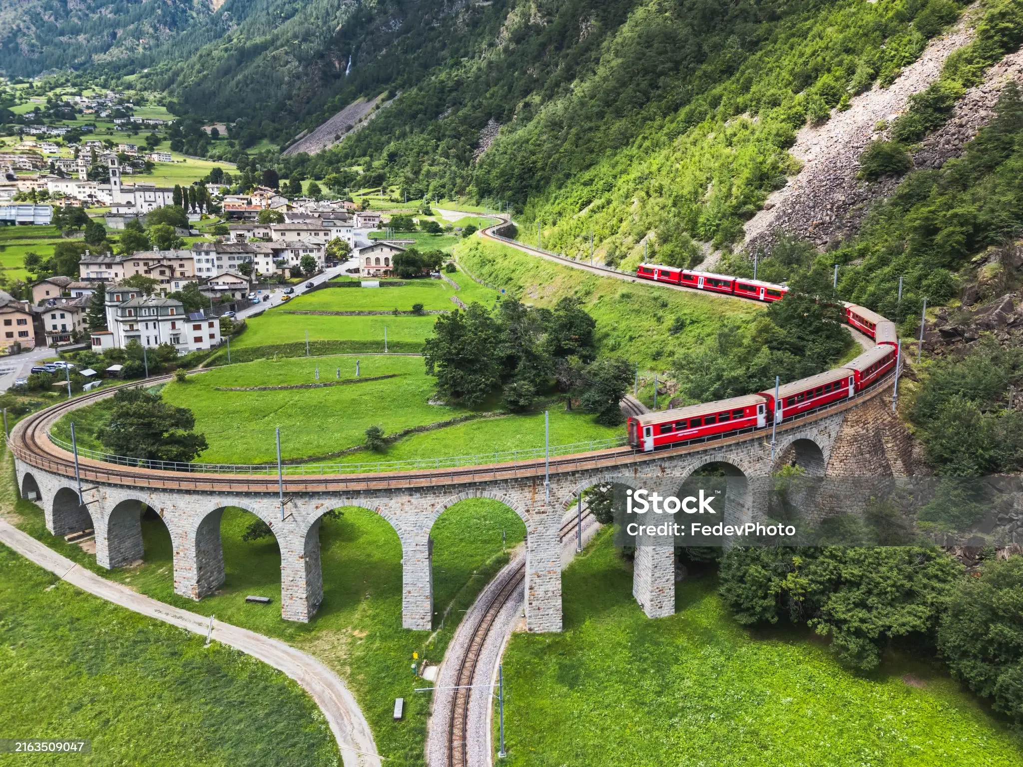 A red train traveling on a curved stone bridge over green fields with a hillside town and mountains in the background.