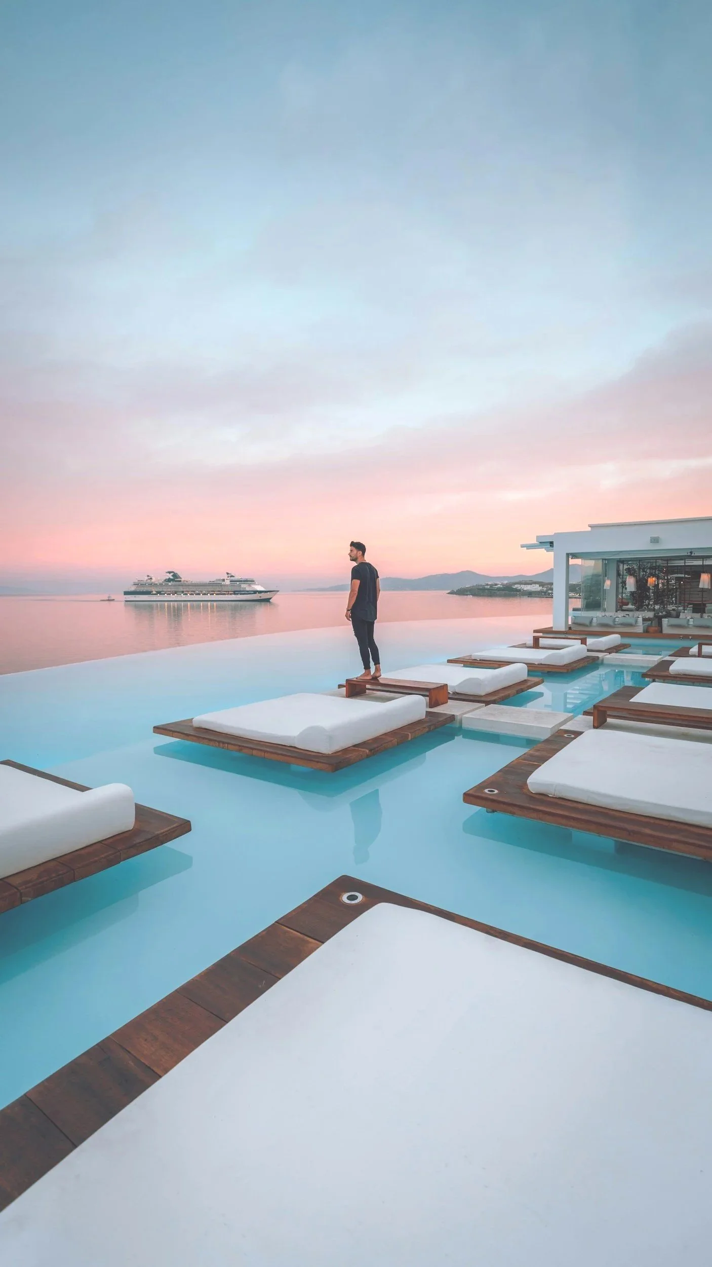 A man standing on a bed in an infinity pool overlooking the ocean during sunset, with a cruise ship in the distance and a modern building on the right.