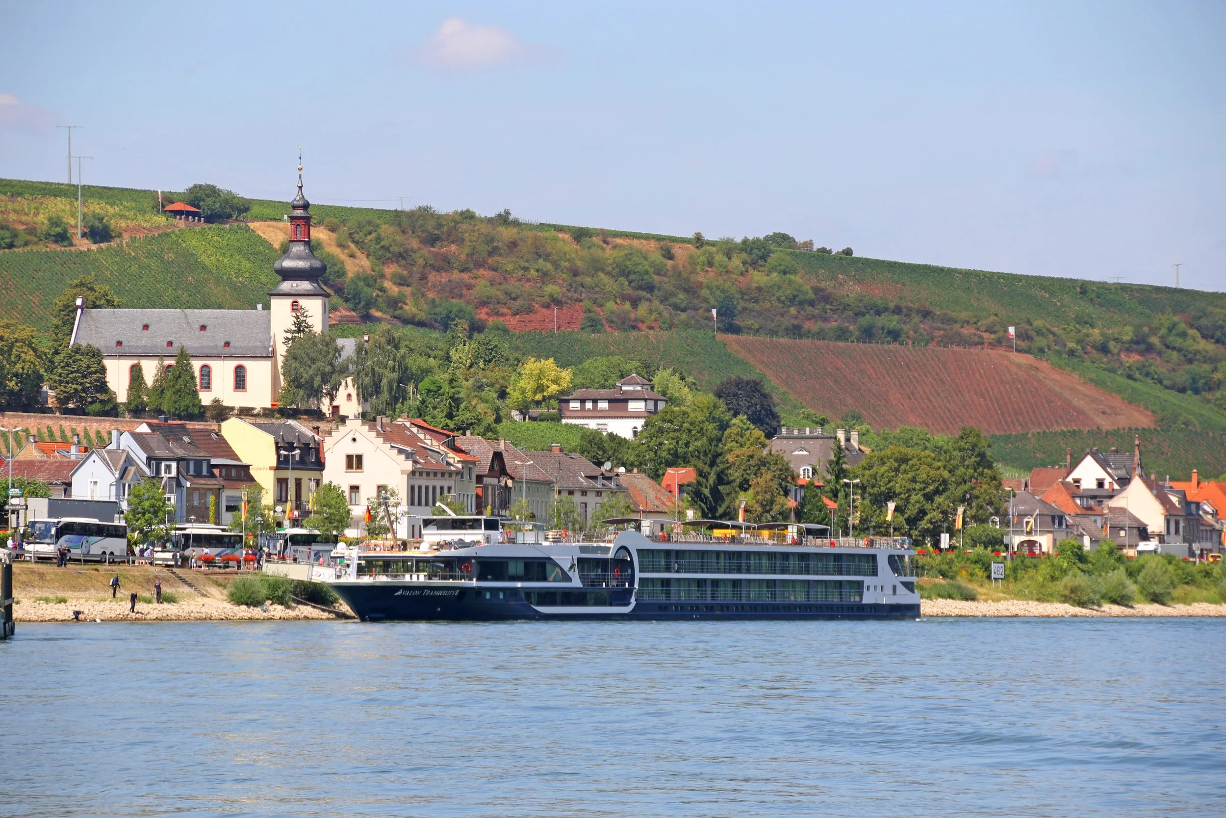 A scenic riverside town with colorful houses, a church with a black steeple, and a large modern cruise ship docked on calm water with vineyards and rolling hills in the background.