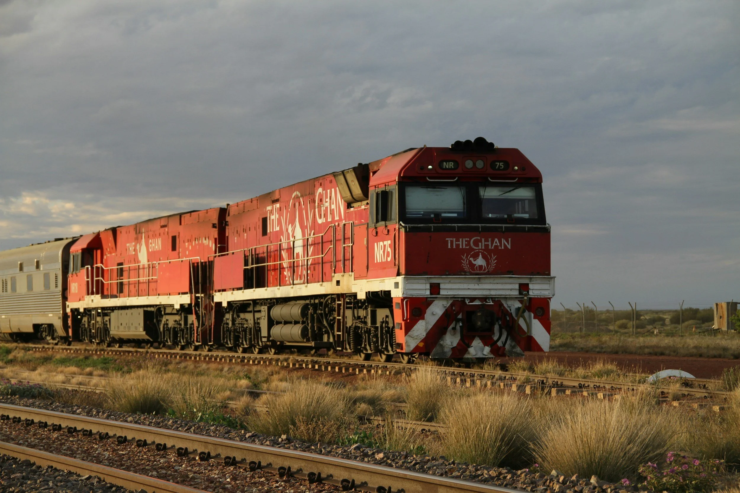 A red locomotive train traveling on railway tracks in an open landscape during daytime with cloudy sky.