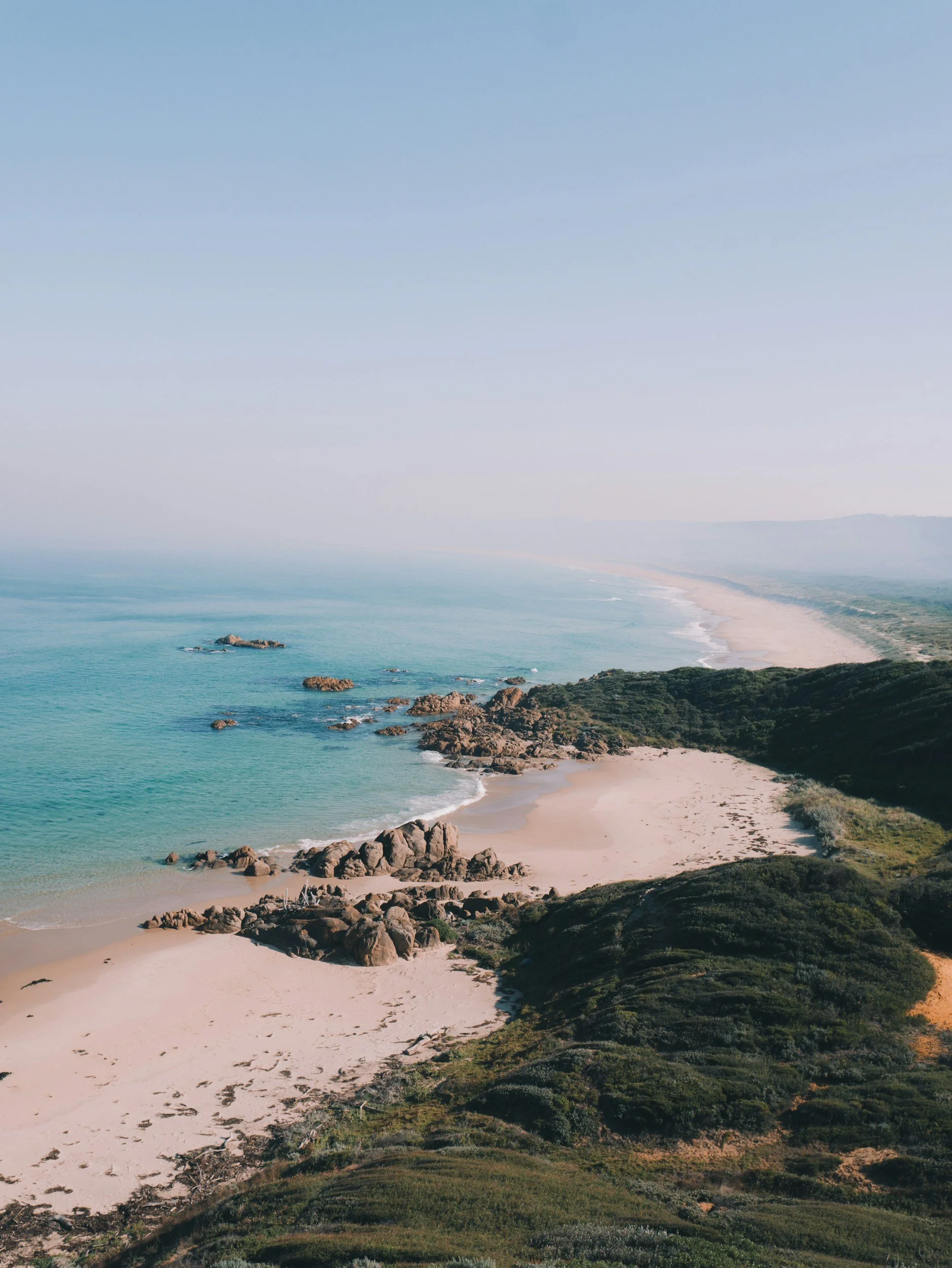 Aerial view of a coastline with sandy beach, rocks, and green hills, along with the ocean extending to the horizon under a clear sky.