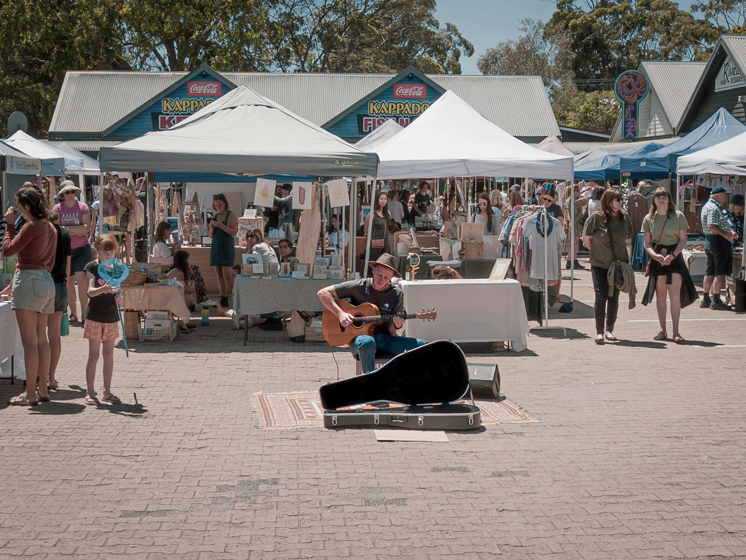 A street market with tents and stalls, a man playing guitar on a rug, and people browsing and walking around on a sunny day.