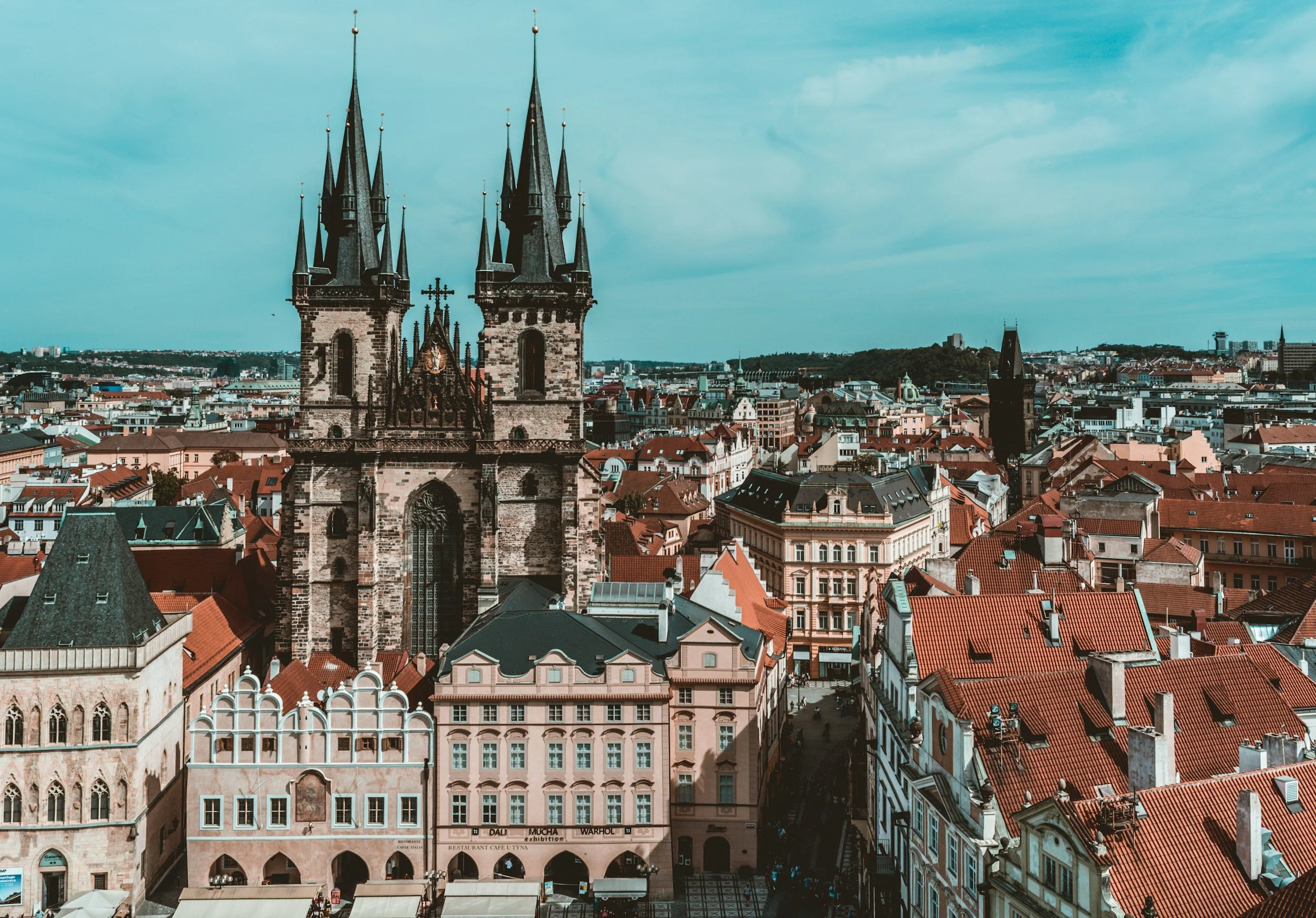 A cityscape featuring a historic church with tall, pointed spires and dense red-tiled rooftops surrounding it, under a partly cloudy sky