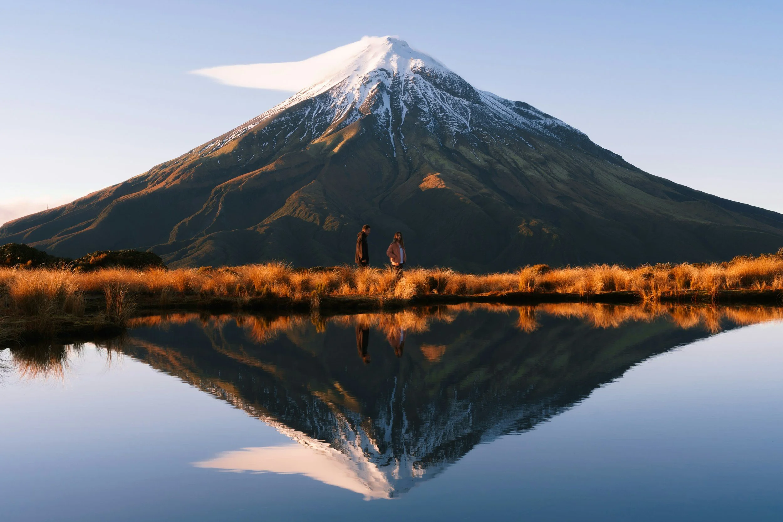 Two people stand on the grassy shore of a lake with a mountain in the background, its peak snow-capped and reflected in the water.