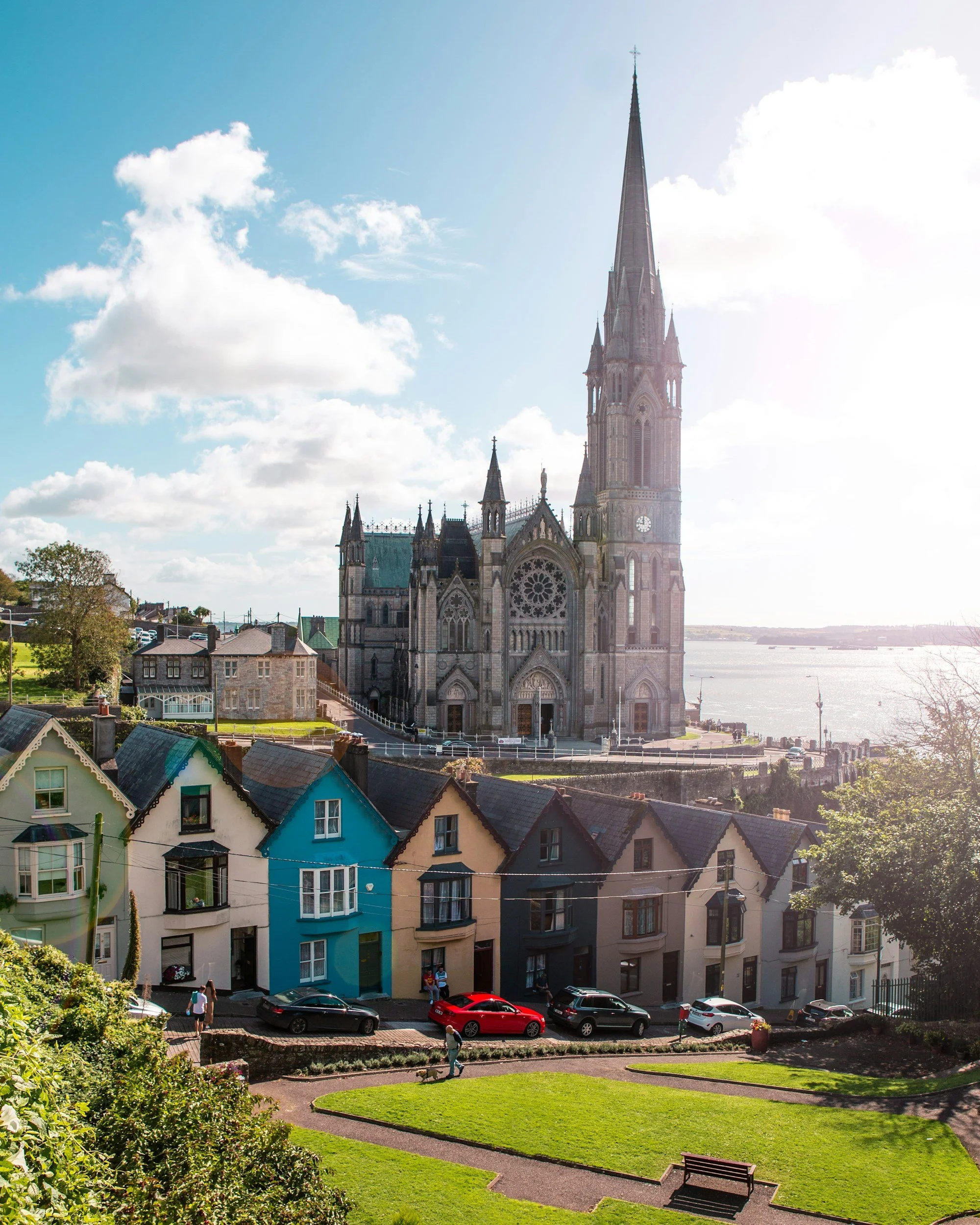 Colorful row houses in front of a large Gothic-style cathedral overlooking a body of water with a park and people walking.