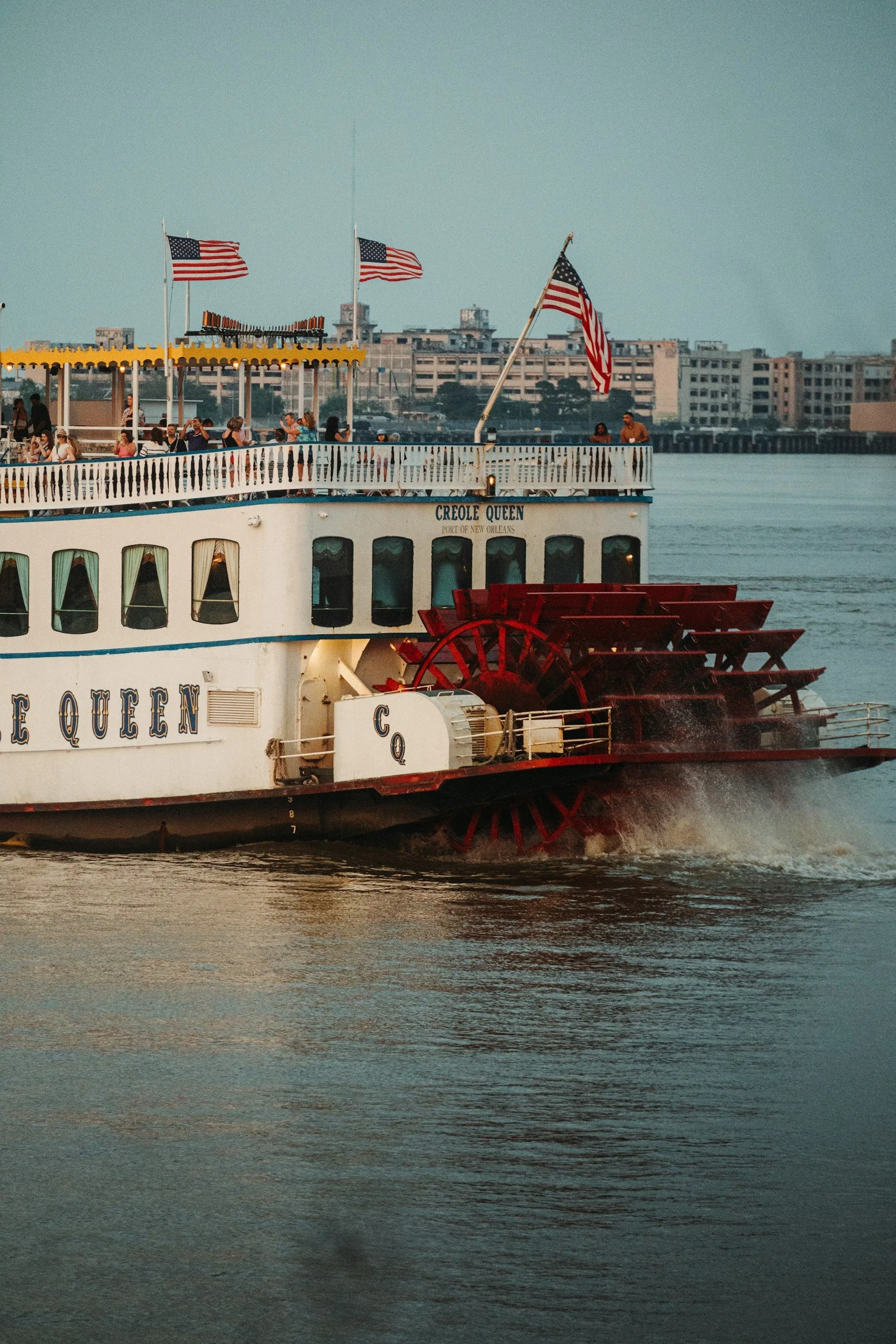 A riverboat named Creole Queen sailing on the water with American flags flying, and a cityscape in the background.