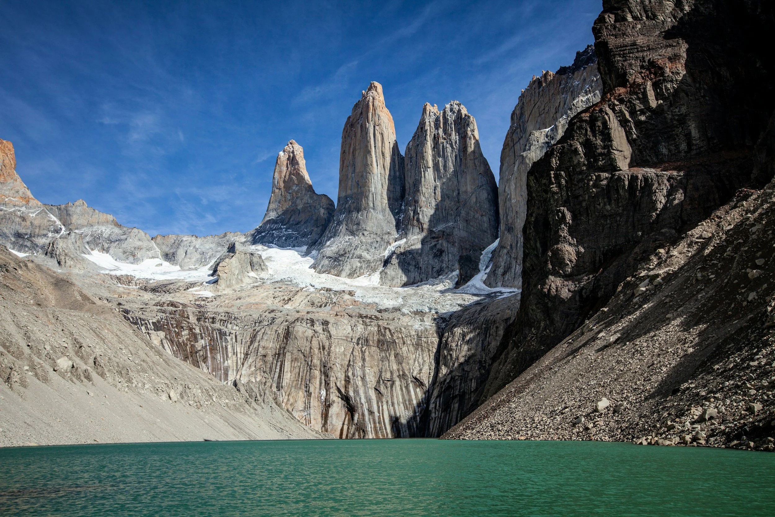 A scenic view of tall mountains with snow patches and a glacier, behind a calm green lake, under a clear blue sky.