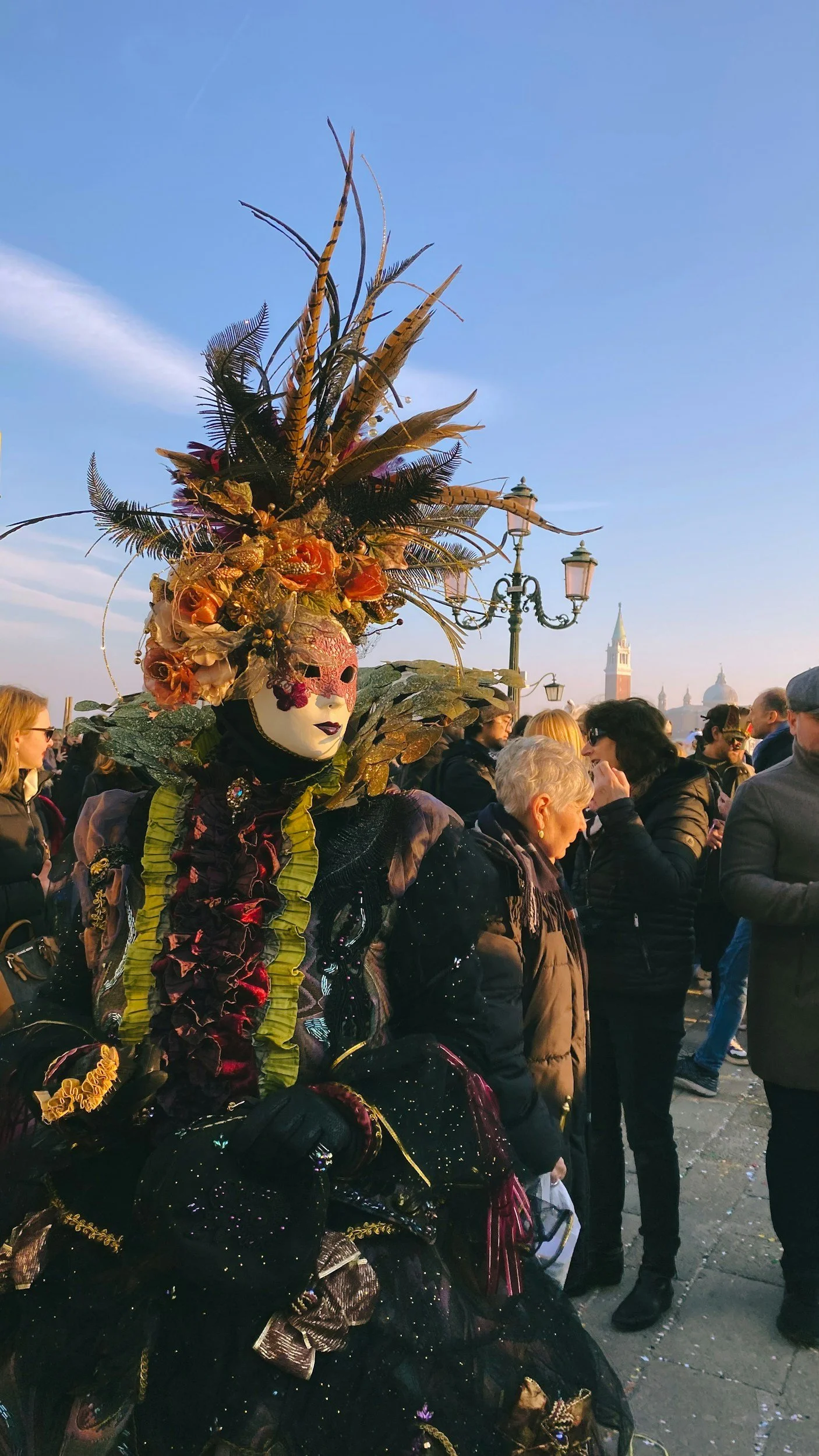 A person dressed in a Venetian-style costume with a elaborate mask and a large headpiece adorned with feathers and flowers, surrounded by a crowd outdoors with historic buildings in the background.