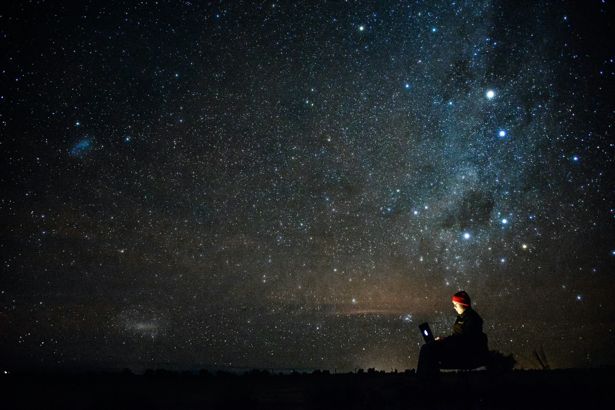 A person sitting on a rock at night, using a laptop, under a starry sky with the Milky Way galaxy visible.