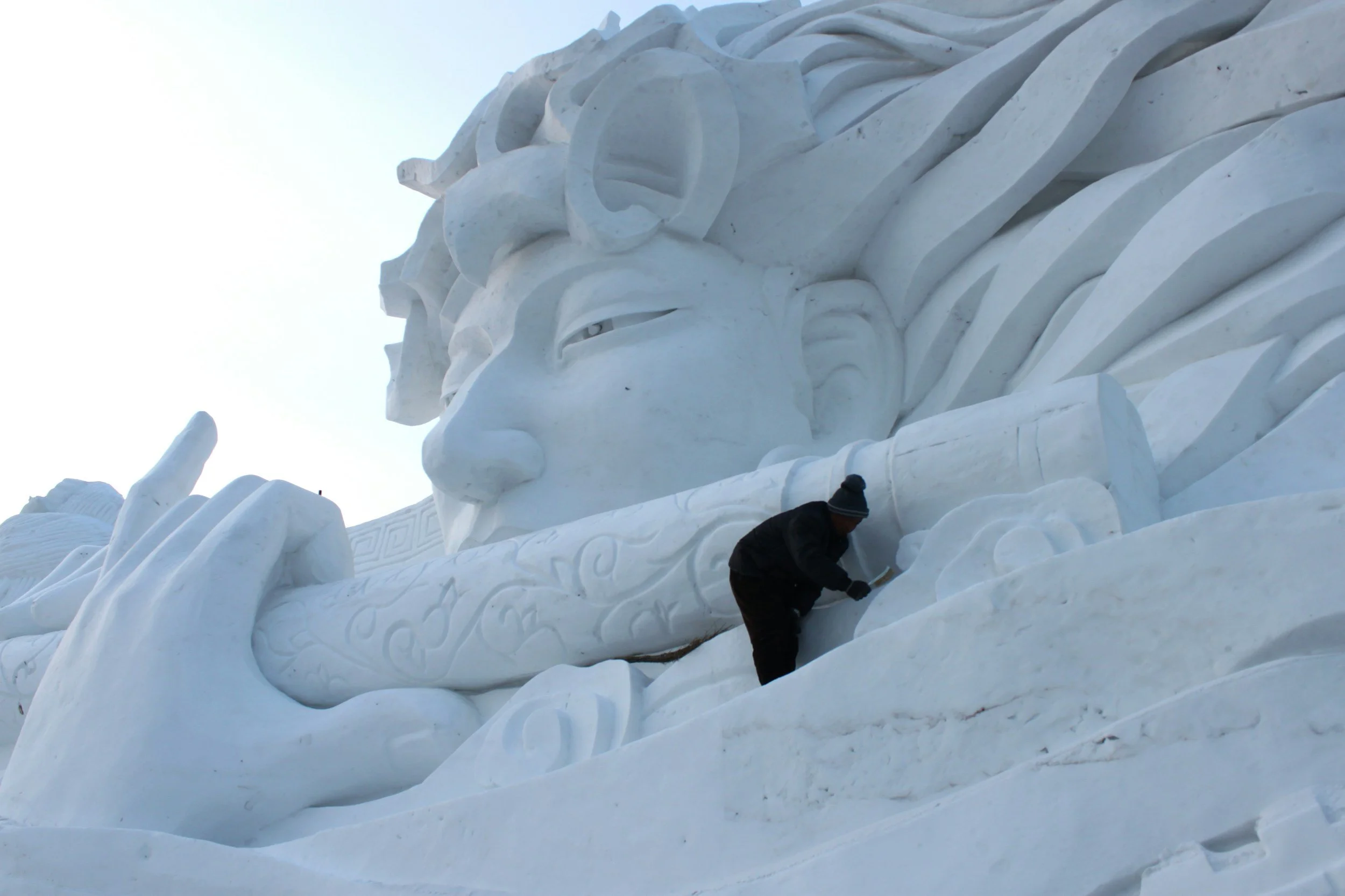 A person working on carving or cleaning a large snow sculpture of a face with long hair and a hand holding an object in front of it.