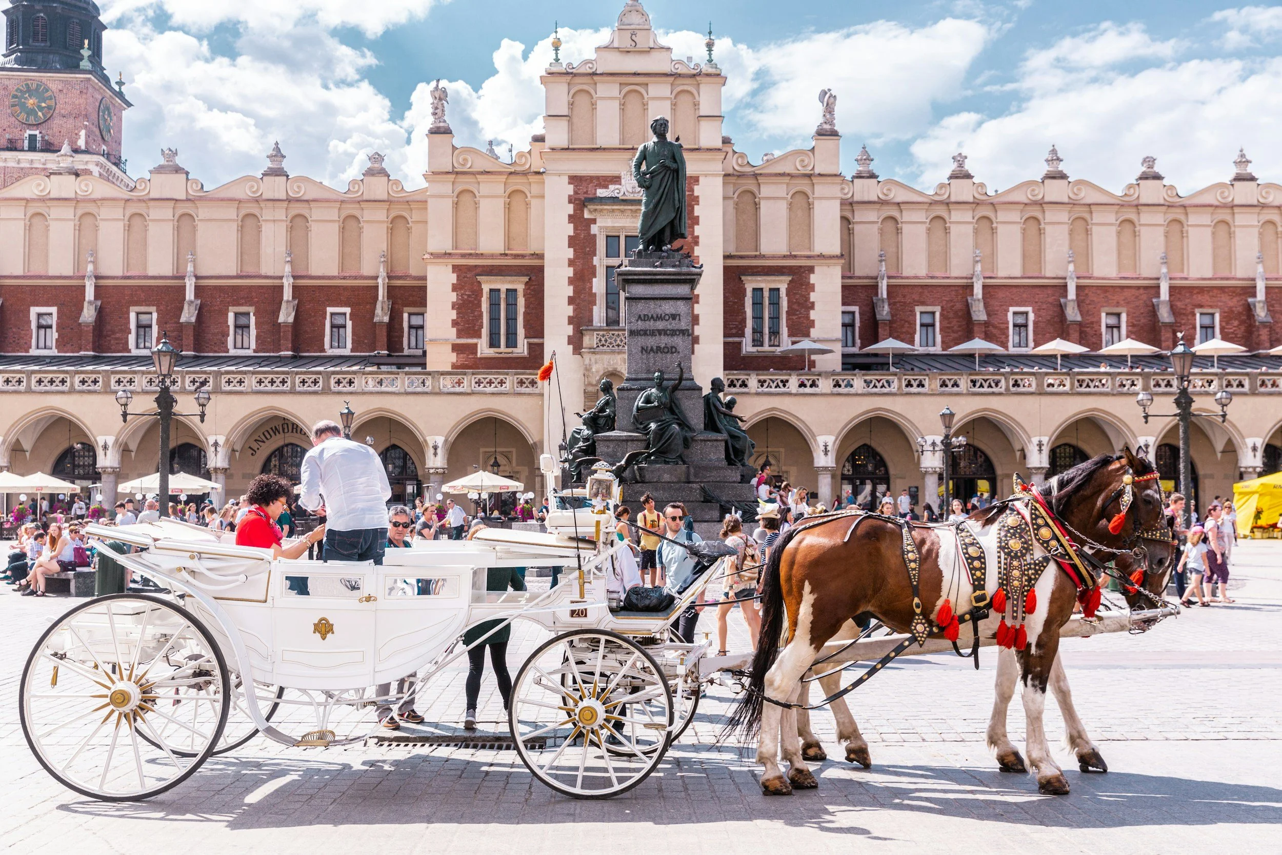A horse-drawn carriage with white body and golden accents parked on a busy town square in front of a historic building with a statue in the center, surrounded by many pedestrians.
