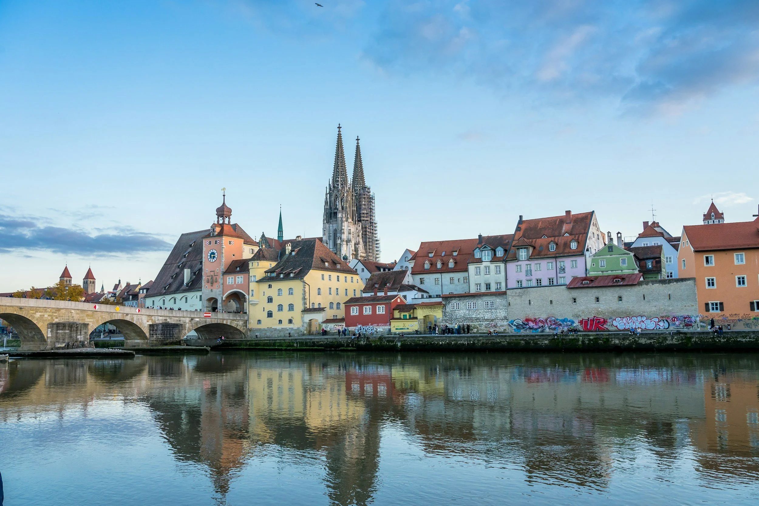 Colorful buildings and a bridge along a river in a European city, with a cathedral featuring tall spires in the background.