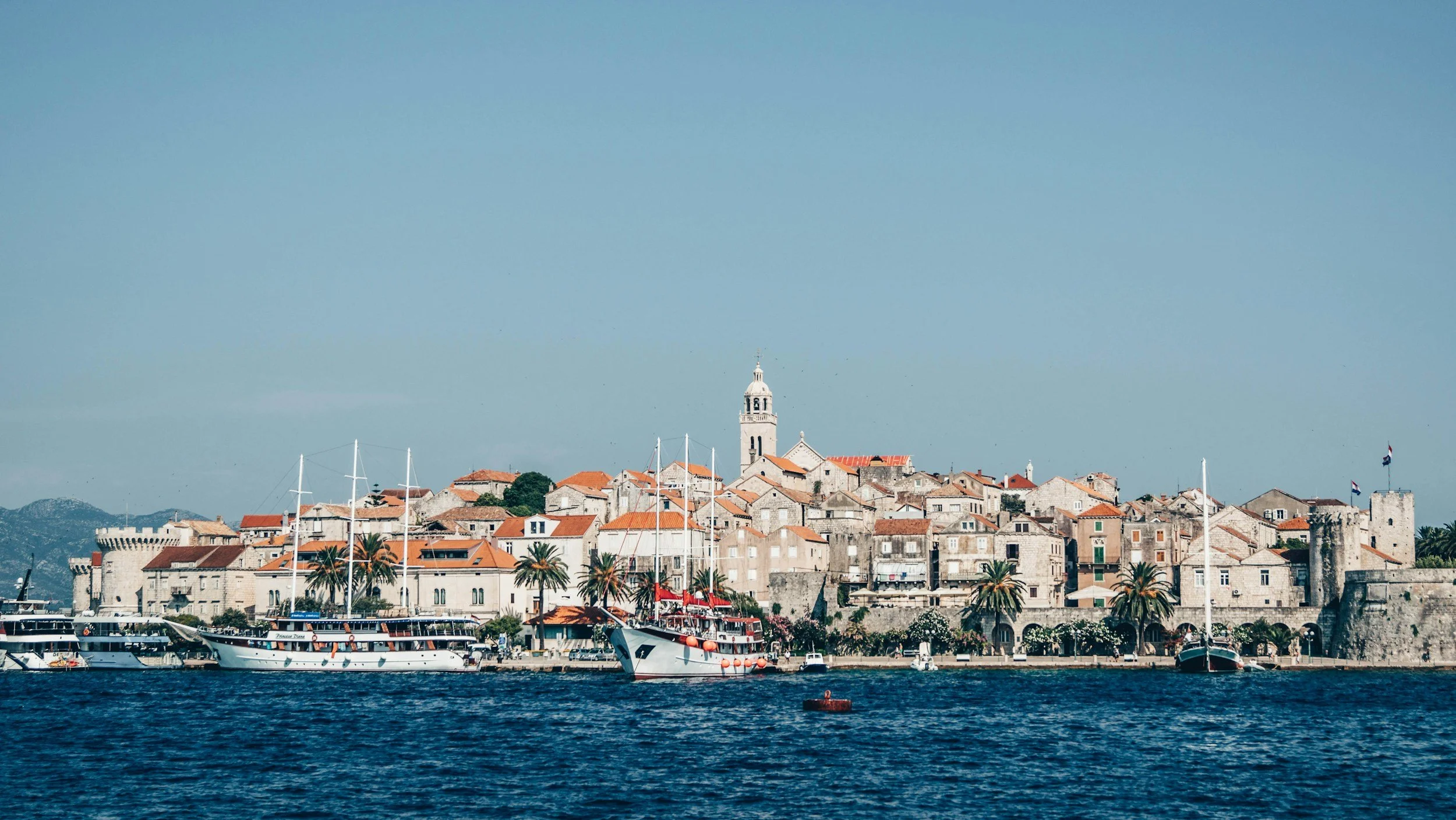 Scenic view of a historic coastal city with stone buildings, towers, and a church atop a hill, along a waterfront with yachts and palm trees under a clear blue sky.
