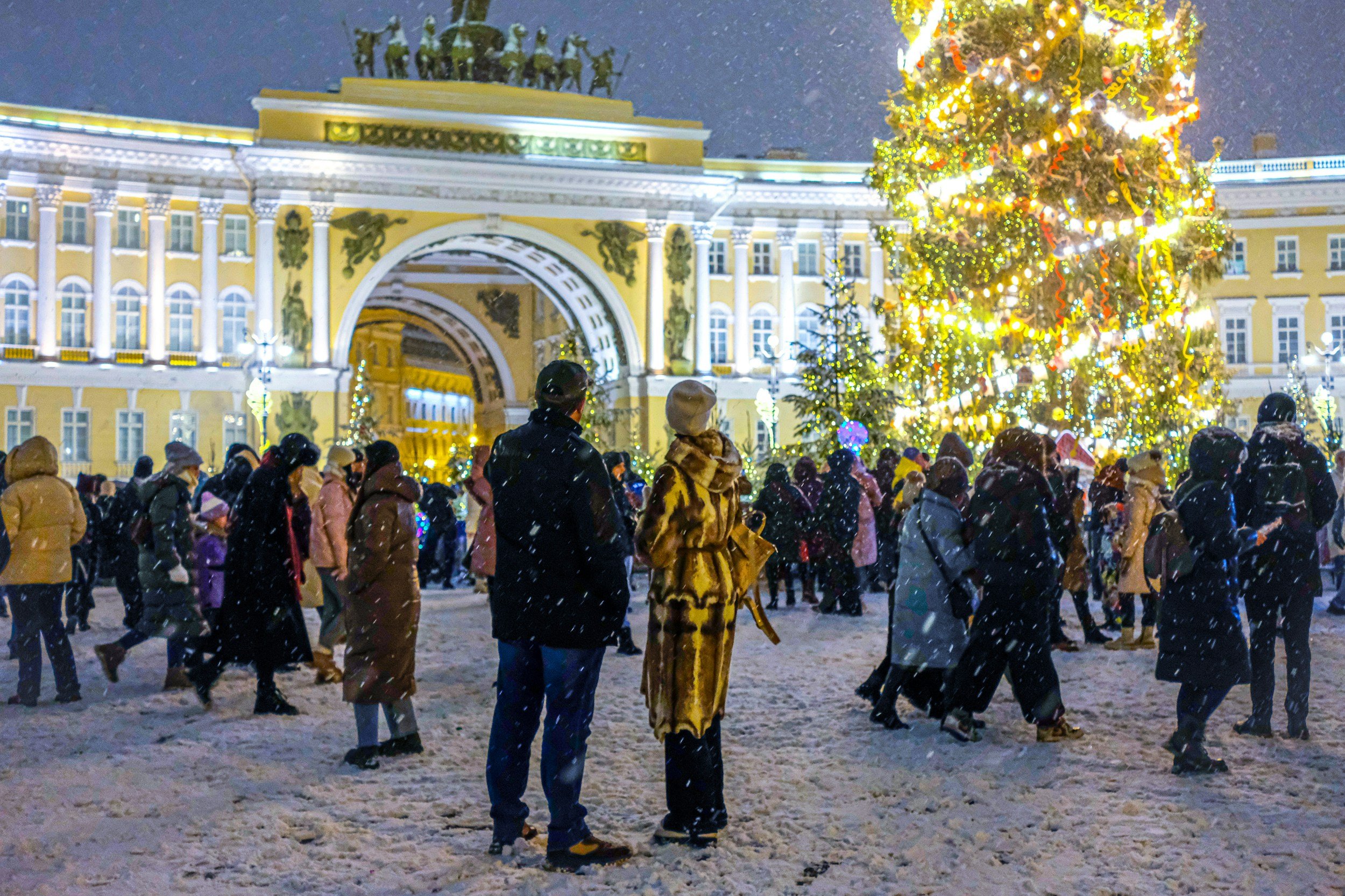 People gathering outside at night for a Christmas celebration, with a large lit Christmas tree and an illuminated yellow building with an arch in the background, snow falling on the ground.