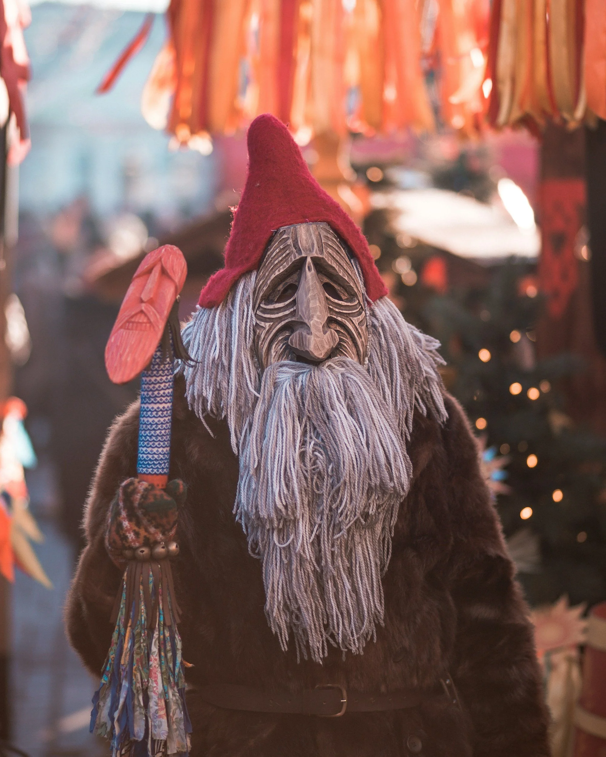 Person dressed in a costume wearing a carved wooden mask, a red hat, a brown fur coat, and patterned gloves, holding a colorful staff. The background features hanging decorations and Christmas lights.