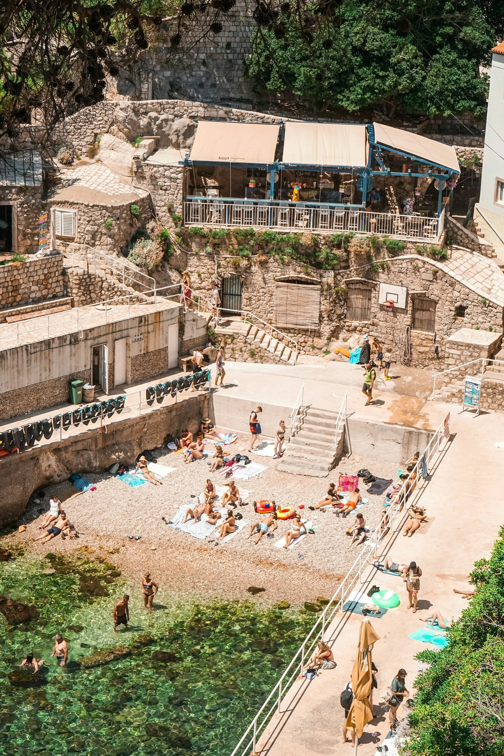 People relaxing and sunbathing on a pebble beach next to a small, clear, green body of water, with some people in the water. There are several towels, beach chairs, and inflatable floaties. Some people are walking along the shoreline and stairs leading to various levels of a rocky, stone building and deck area with shaded seating.