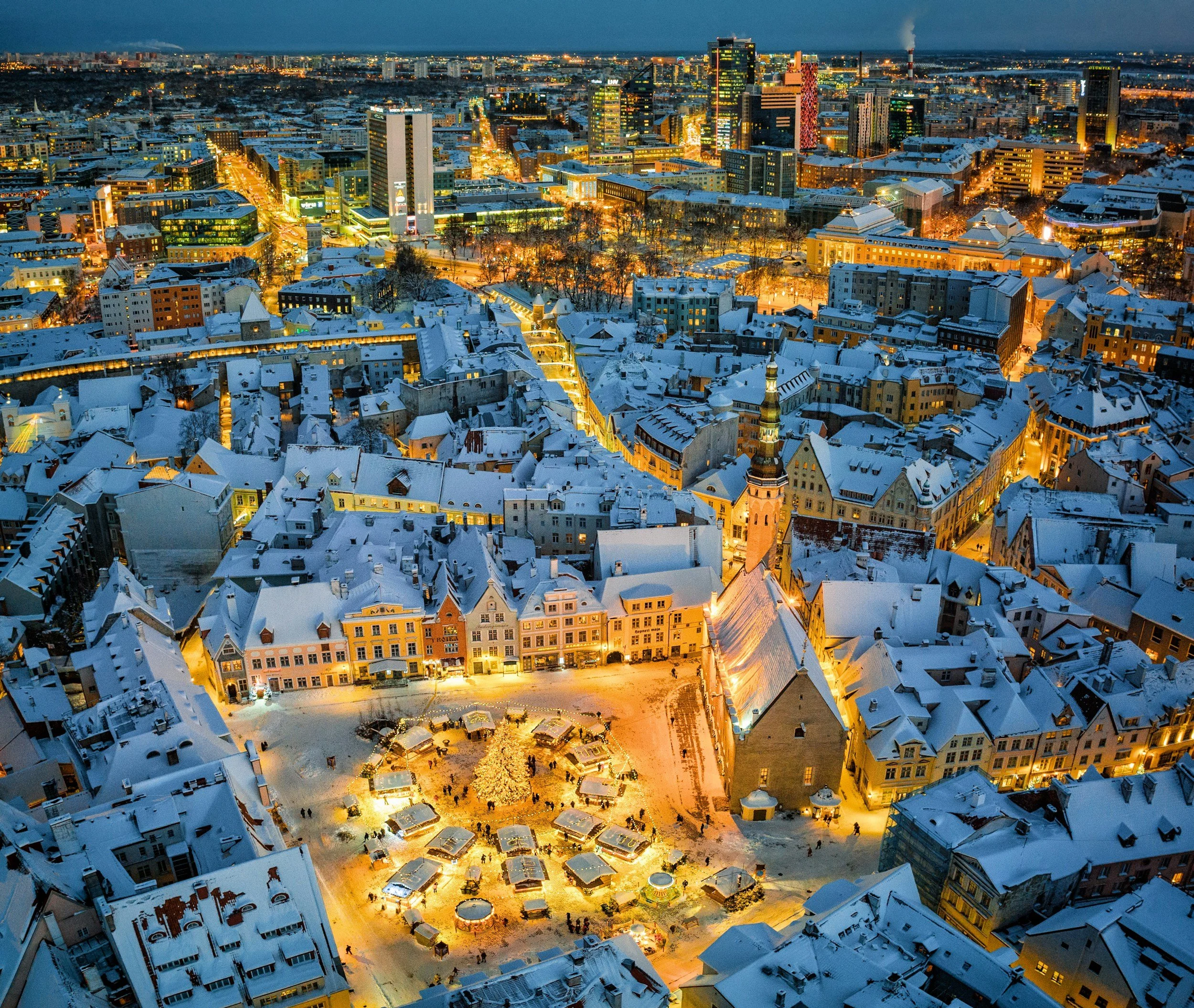 Aerial view of a snow-covered cityscape at dusk, featuring historic buildings, modern skyscrapers, and a festive outdoor market with a decorated Christmas tree.