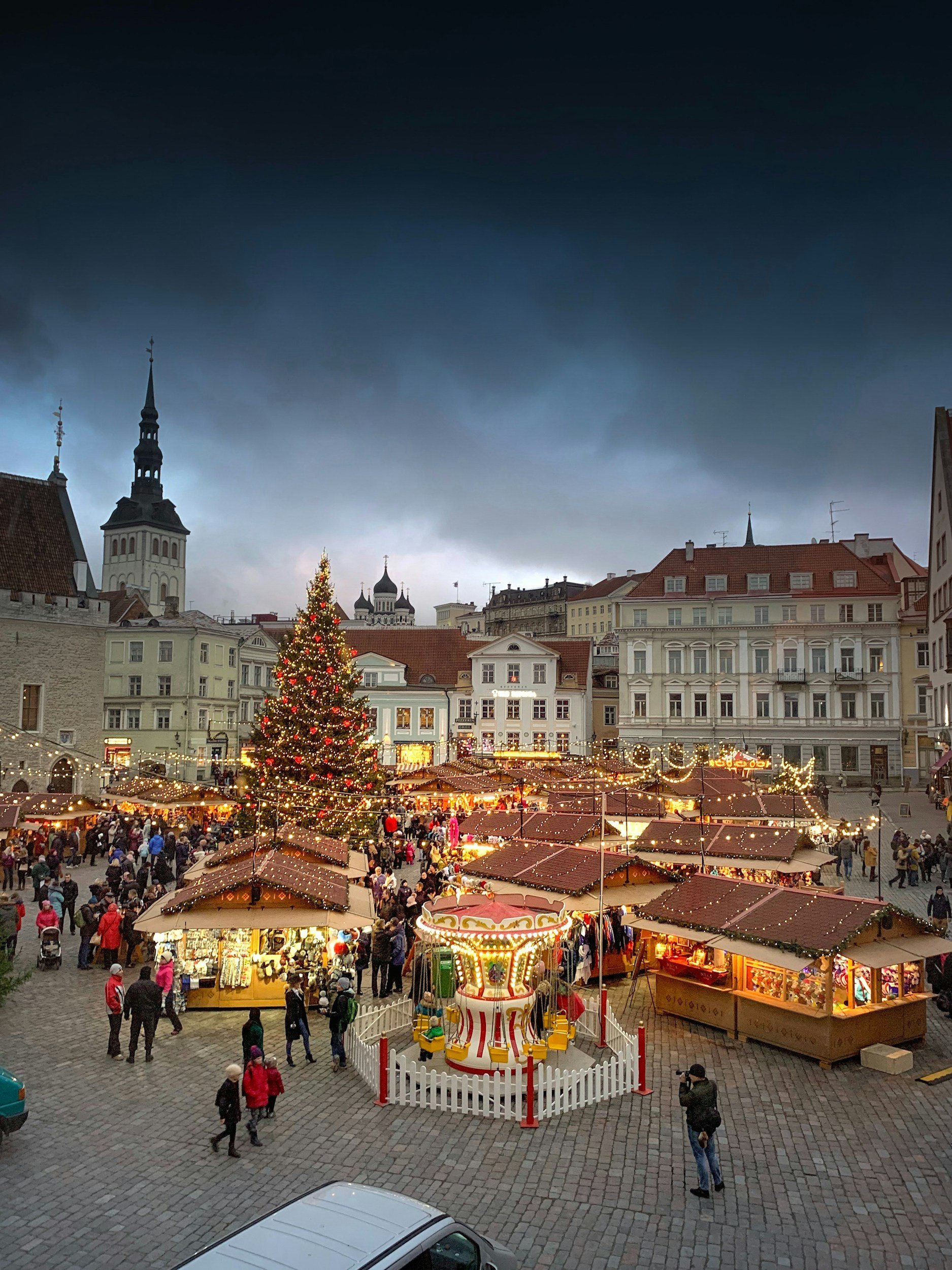A lively Christmas market in a historic European town square at dusk, featuring a decorated Christmas tree, festive stalls with lights, and a carousel at the center, with dark stormy skies overhead.