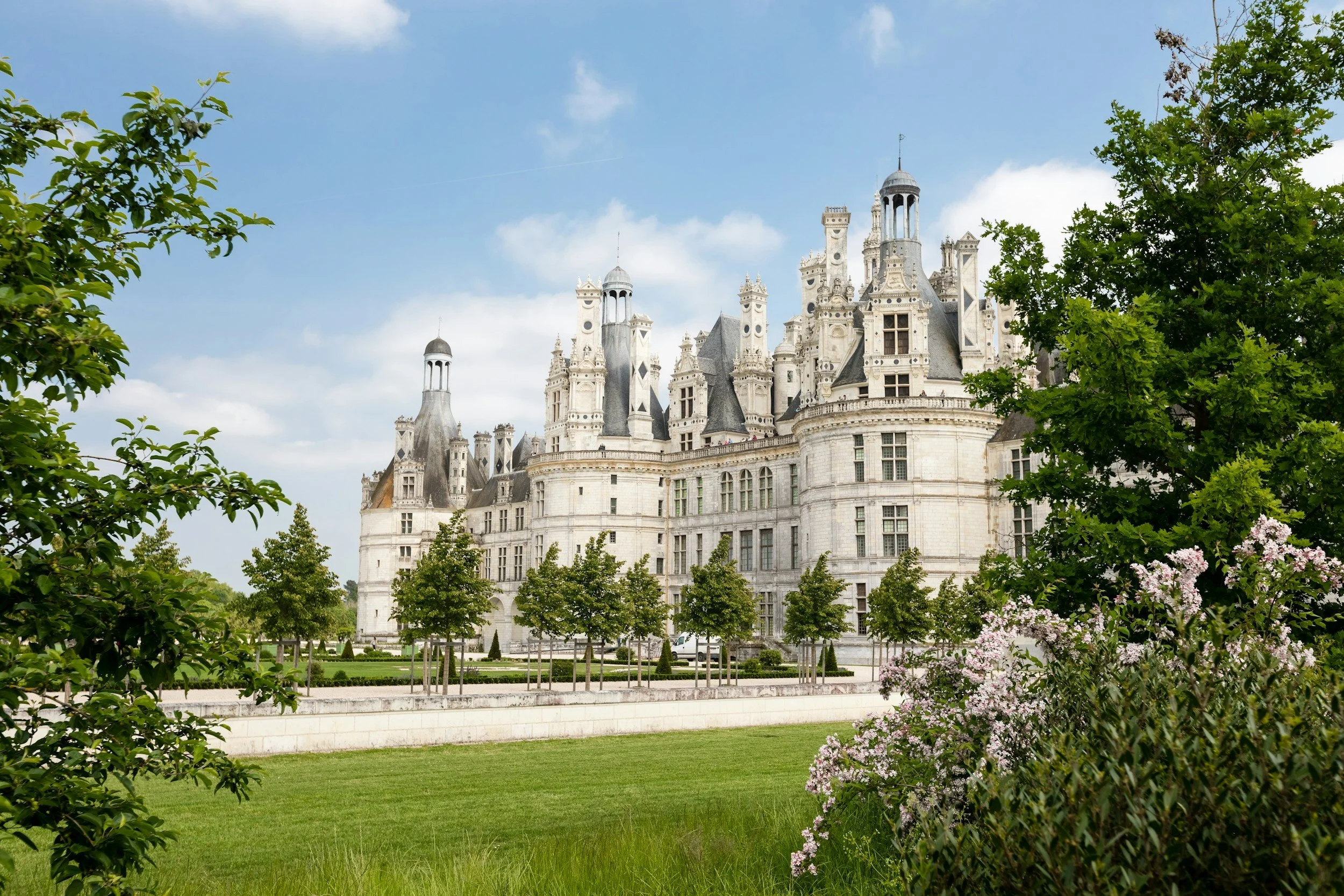 A large, ornate castle with turrets and towers surrounded by green trees and shrubs under a partly cloudy sky.