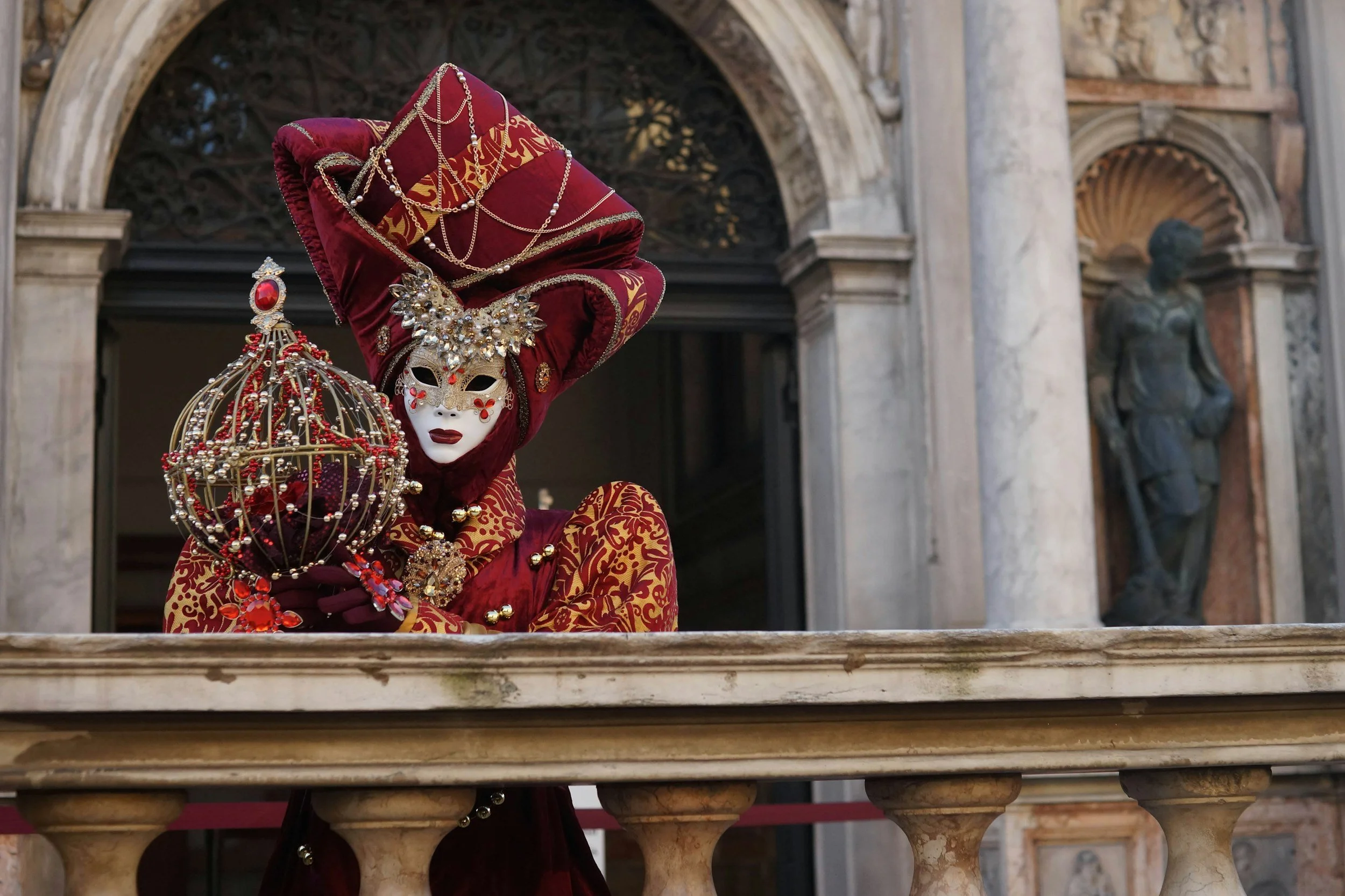 Person wearing an elaborate Venetian carnival costume with a white mask, red and gold embroidered dress, large headdress, and jewelry, standing behind a marble railing in a historic building with classical architecture and statues.