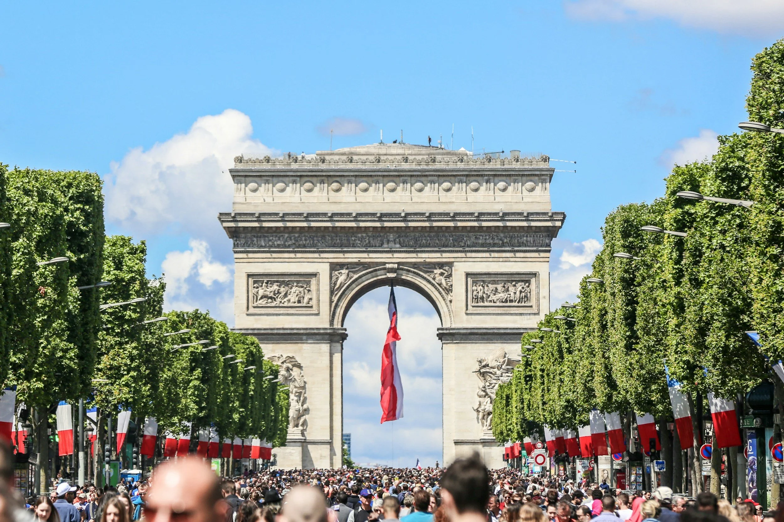 The Arc de Triomphe in Paris, France, decorated with French flags during a large crowd gathering on a sunny day.