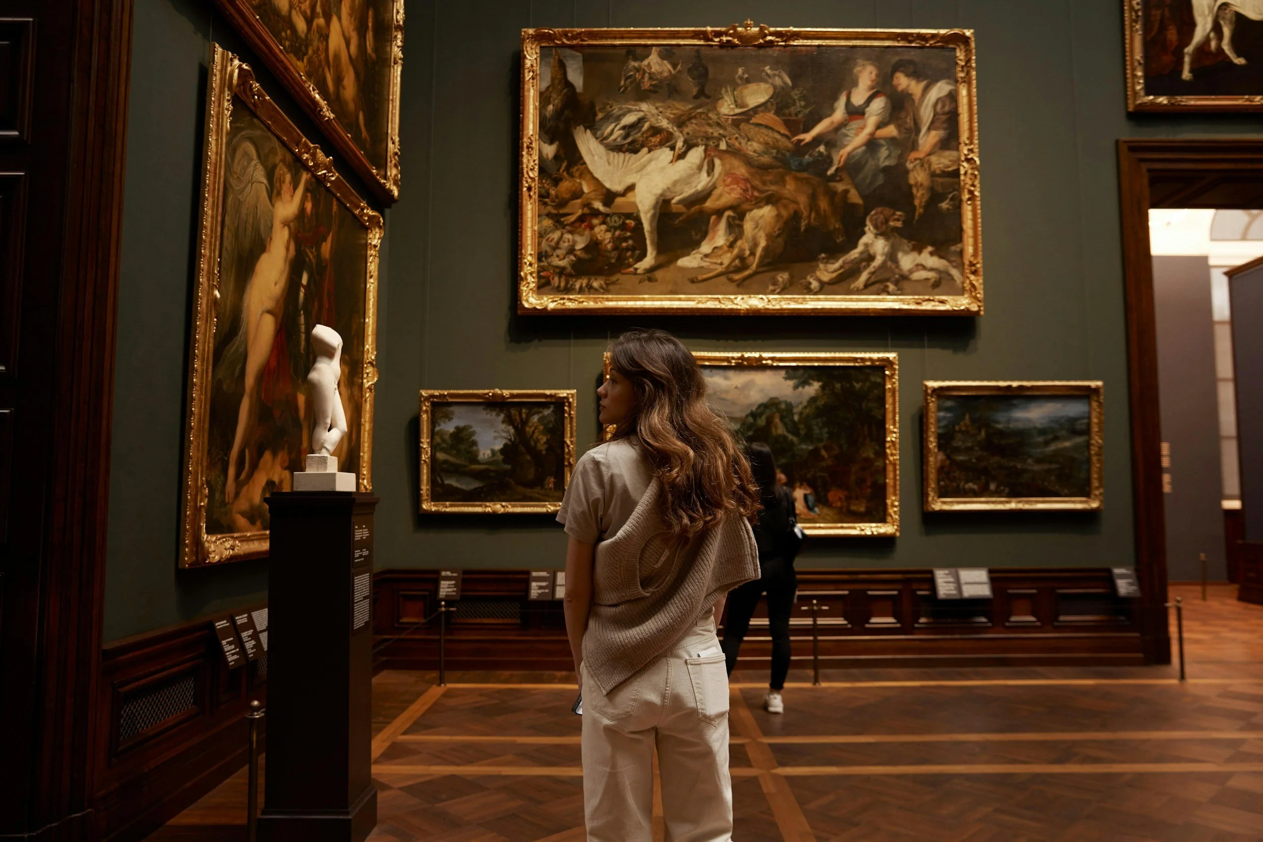 A woman with long wavy hair wearing a beige sweater and white pants looking at paintings and sculptures in an art gallery.