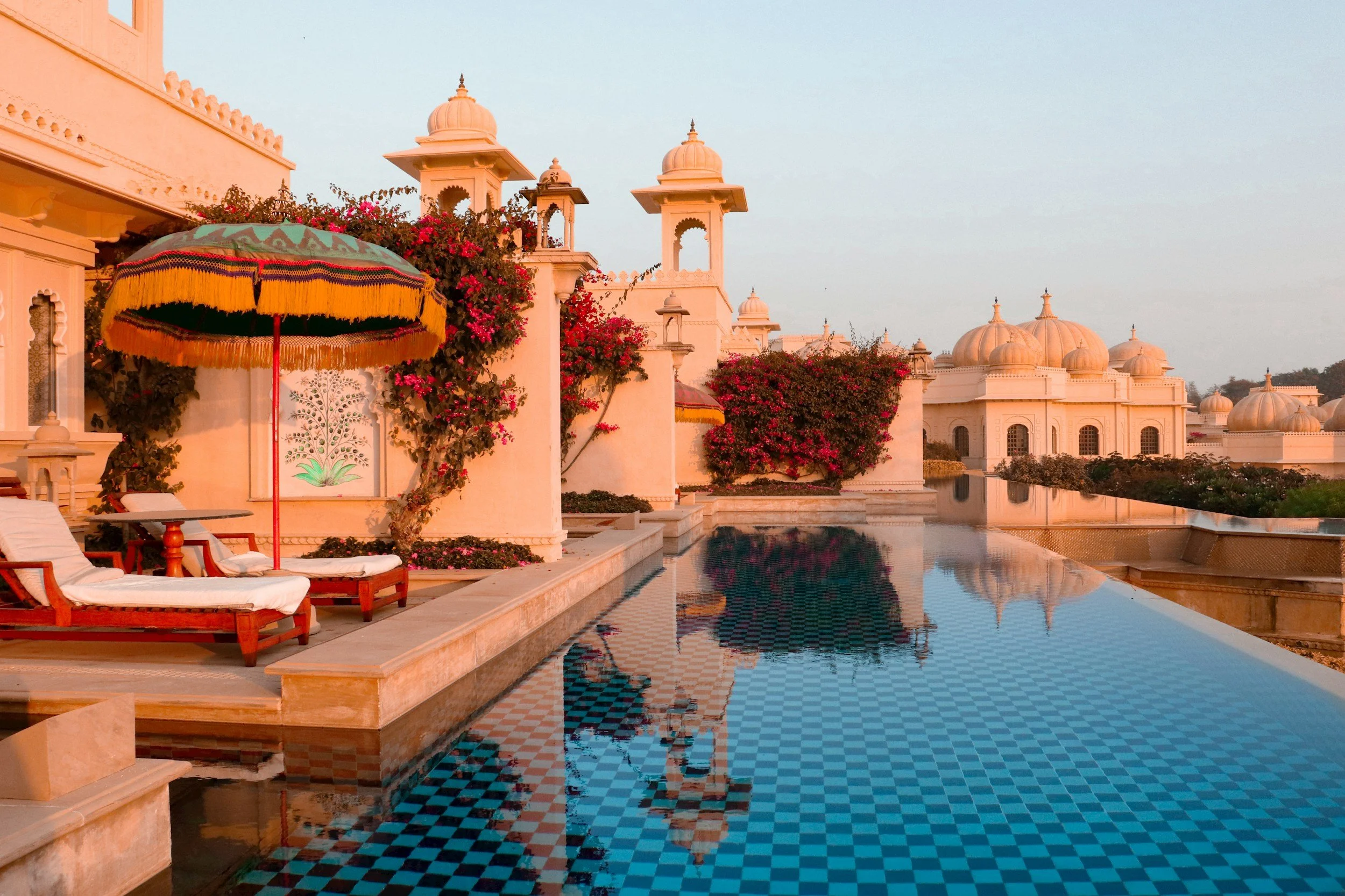 A luxurious poolside scene at sunset with white architecture, domed structures, lush pink flowering bushes, and a colorful umbrella with lounge chairs nearby.