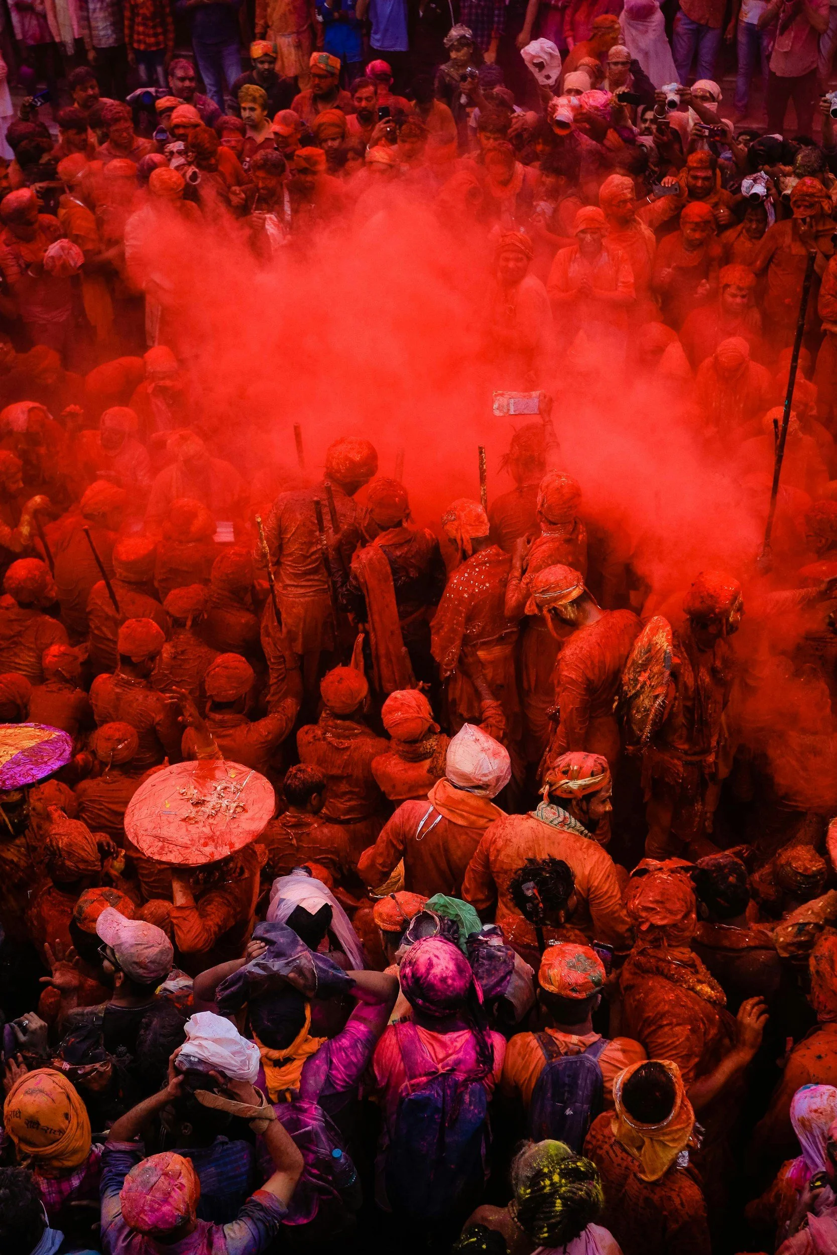 A large crowd celebrating the Holi festival, with people covered in red and pink powder, some holding umbrellas and taking photos, as red powder fills the air.