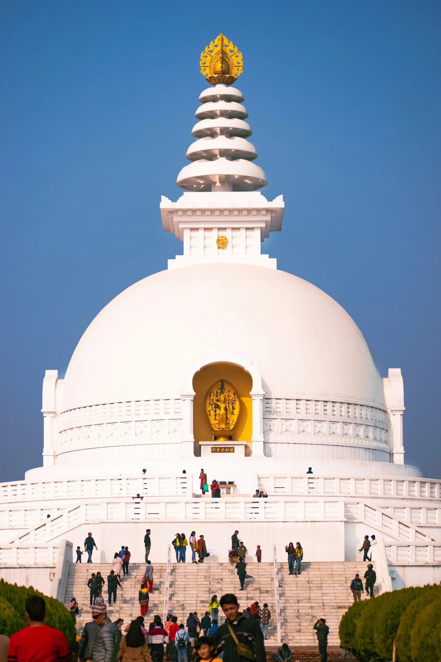 White stupa with a large dome, golden ornament on top, and a central niche containing a gold-colored statue, with people walking up stairs and around the structure.