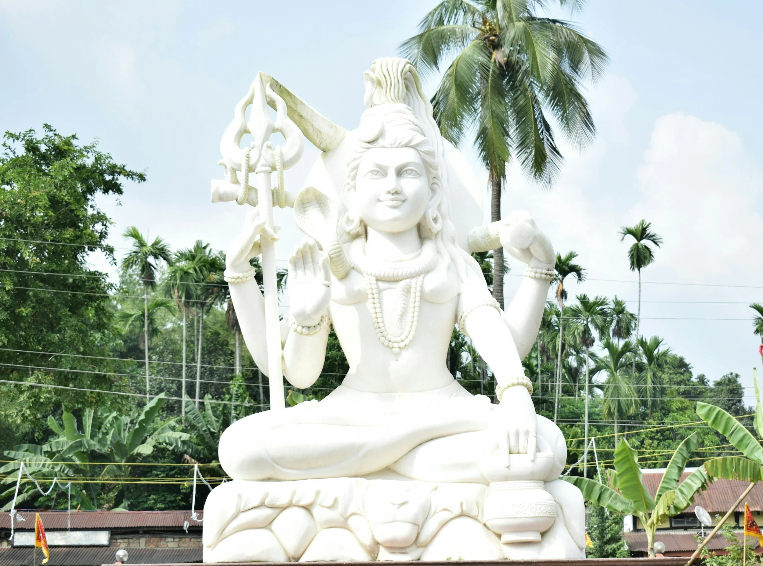 Large white statue of a deity with multiple arms, seated in a cross-legged position outdoors, surrounded by palm trees and tropical plants.