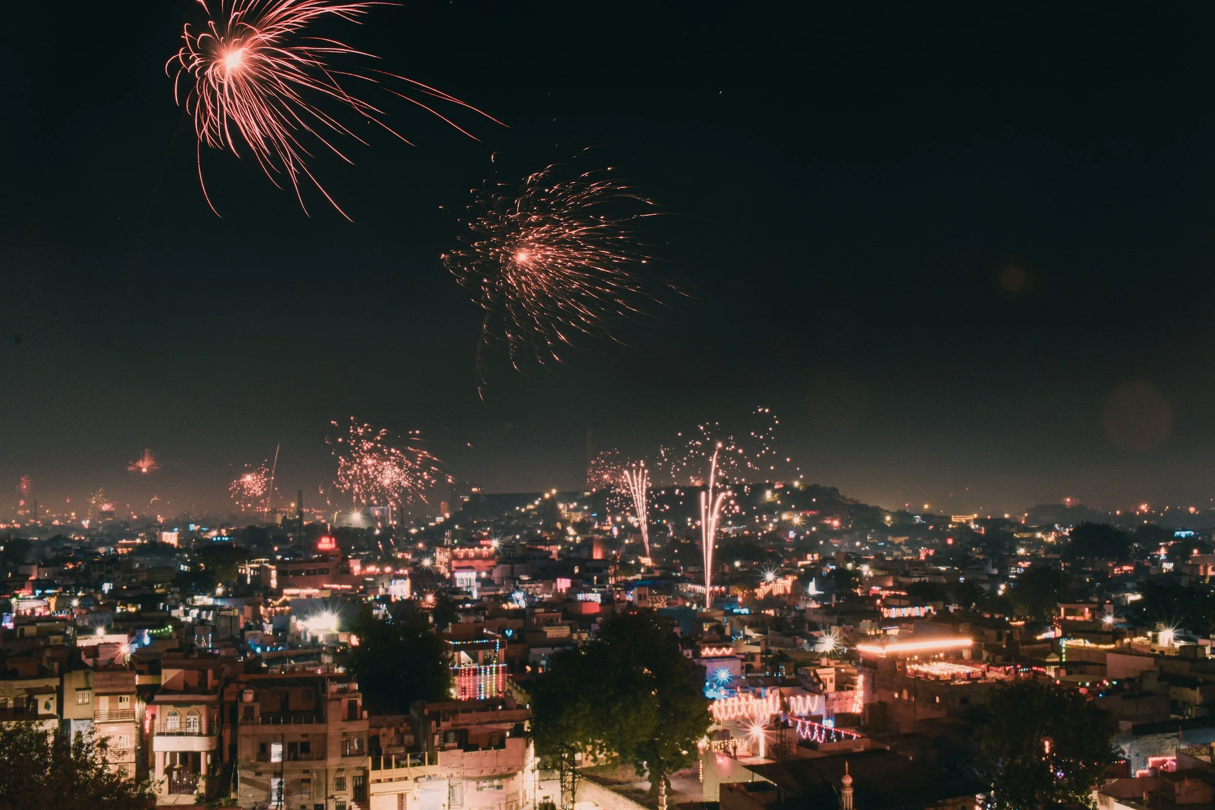 Nighttime cityscape with fireworks exploding in the sky over rooftops decorated with colorful lights.