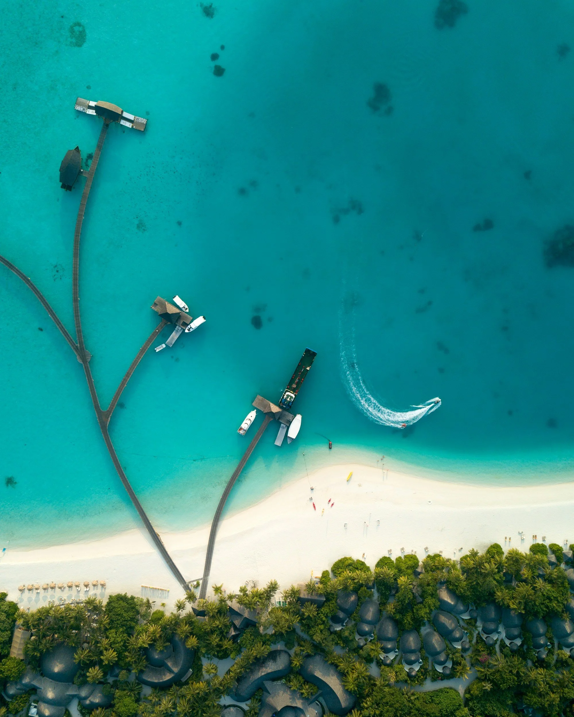 Aerial view of a coastline with a beach, water, and dock structures connected by pathways, with boats and people visible.